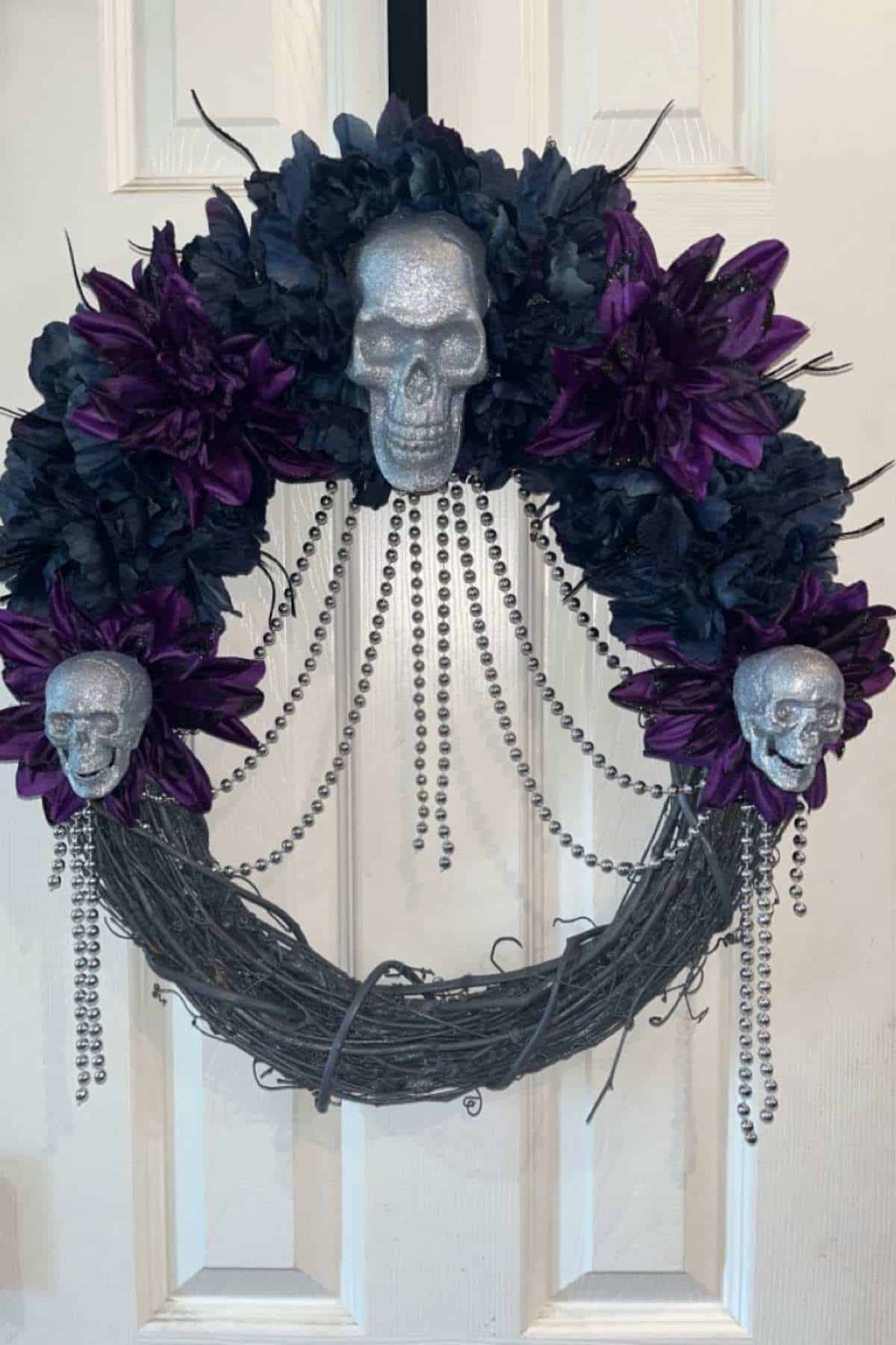A wreath decorated with silver skulls, dark purple flowers, black leaves, and draped silver beads hanging on a white door.