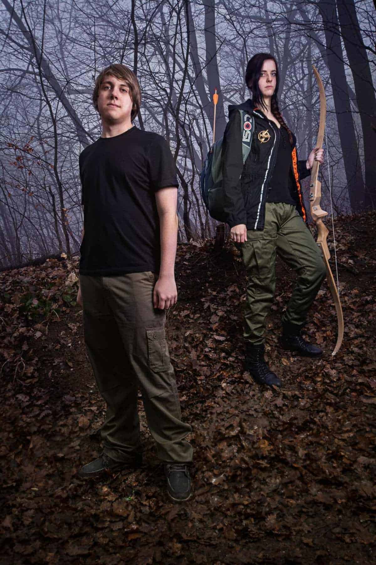 Two people stand in a forest with fallen leaves; one holds a bow and wears a backpack, while the other stands with hands at their sides. The background is foggy and filled with bare trees.