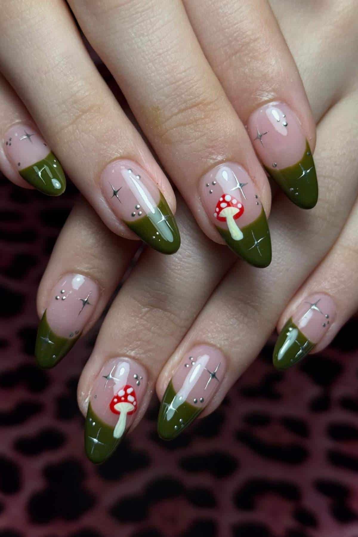 Hands with long, almond-shaped nails featuring green French tips, small silver star accents, and red-and-white mushroom designs on two nails.