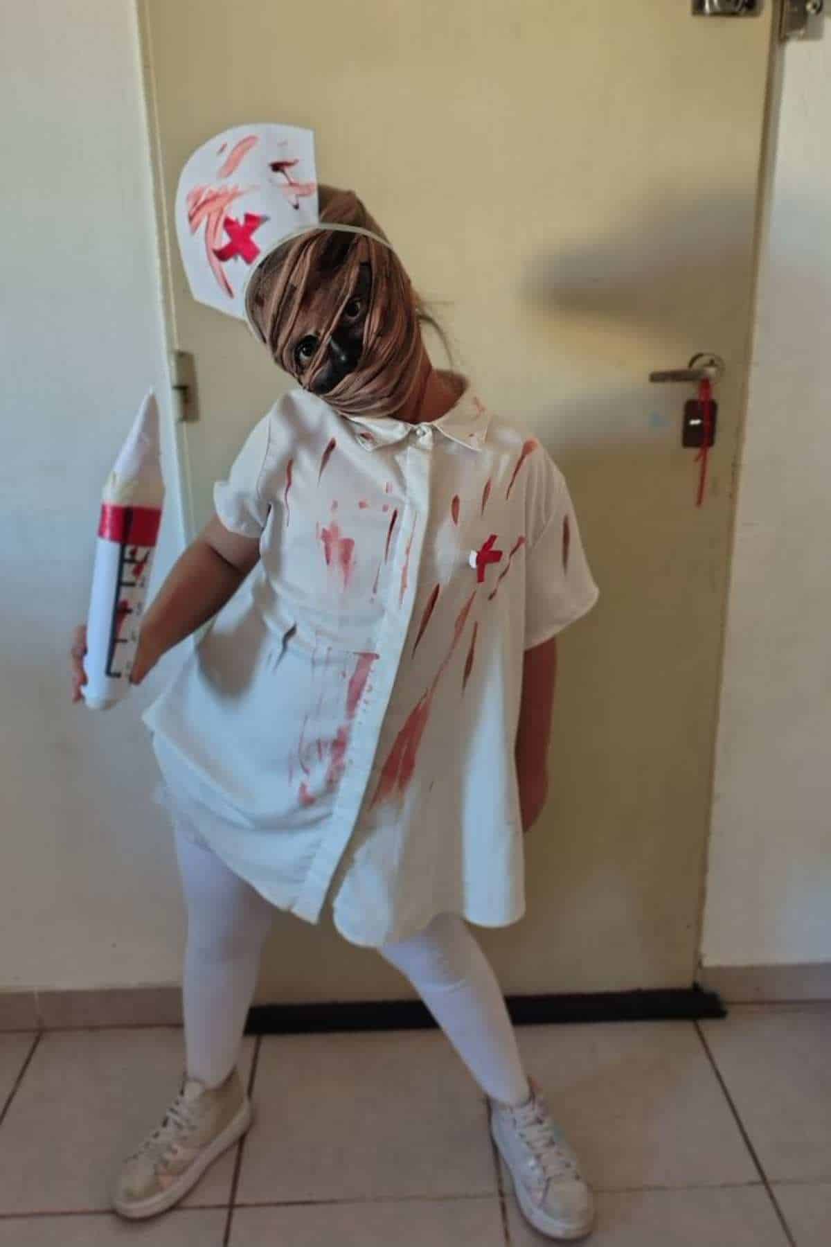 Child in a white nurse costume with red stains, holding a large syringe prop, wearing a nurse hat and a mask wrapped around their face, standing indoors.