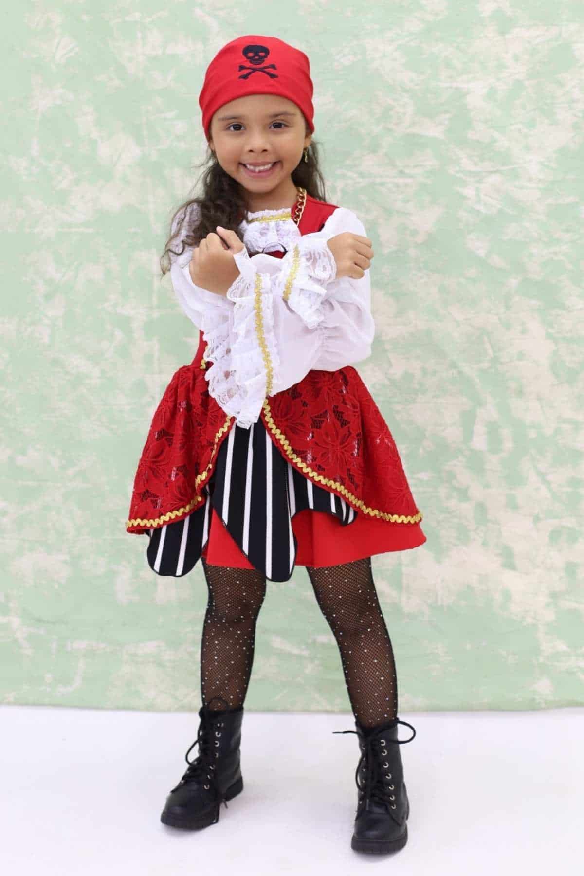 Young girl in a pirate costume with a red headscarf, black boots, and striped skirt stands smiling against a green and white backdrop.