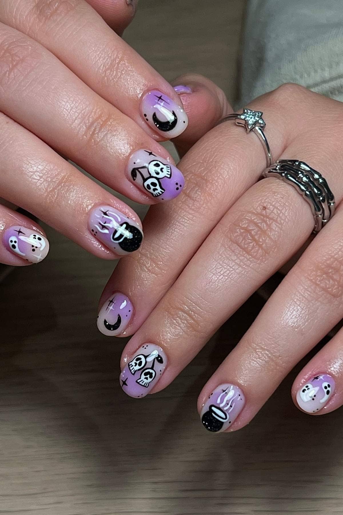 Hands with pastel purple and nude Halloween-themed nail art, featuring ghosts, skulls, crescent moons, and dripping designs. The person wears silver rings, including one with a star shape.