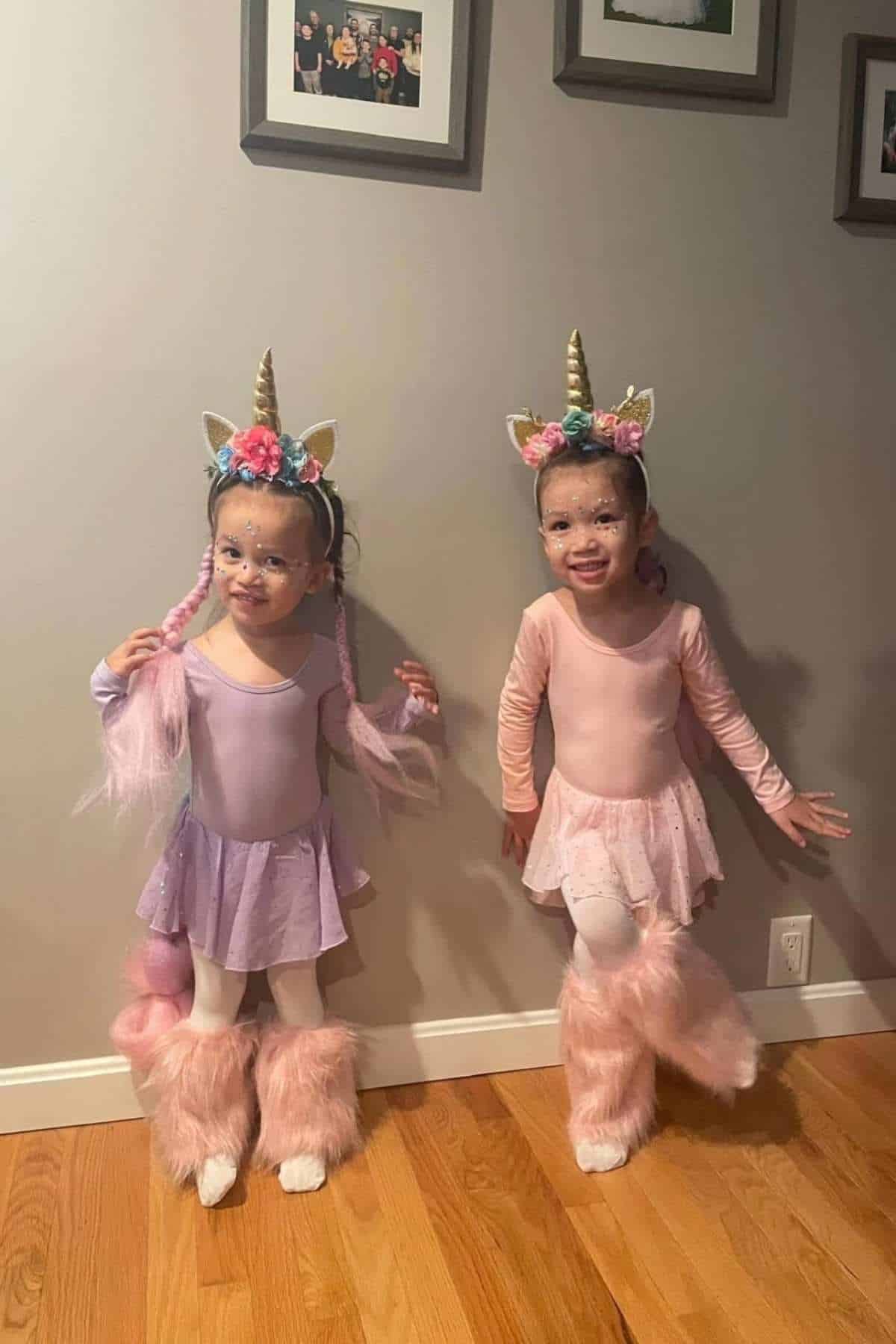 Two young children in pink and purple unicorn costumes stand side by side indoors, smiling at the camera, with framed photos on the wall behind them.