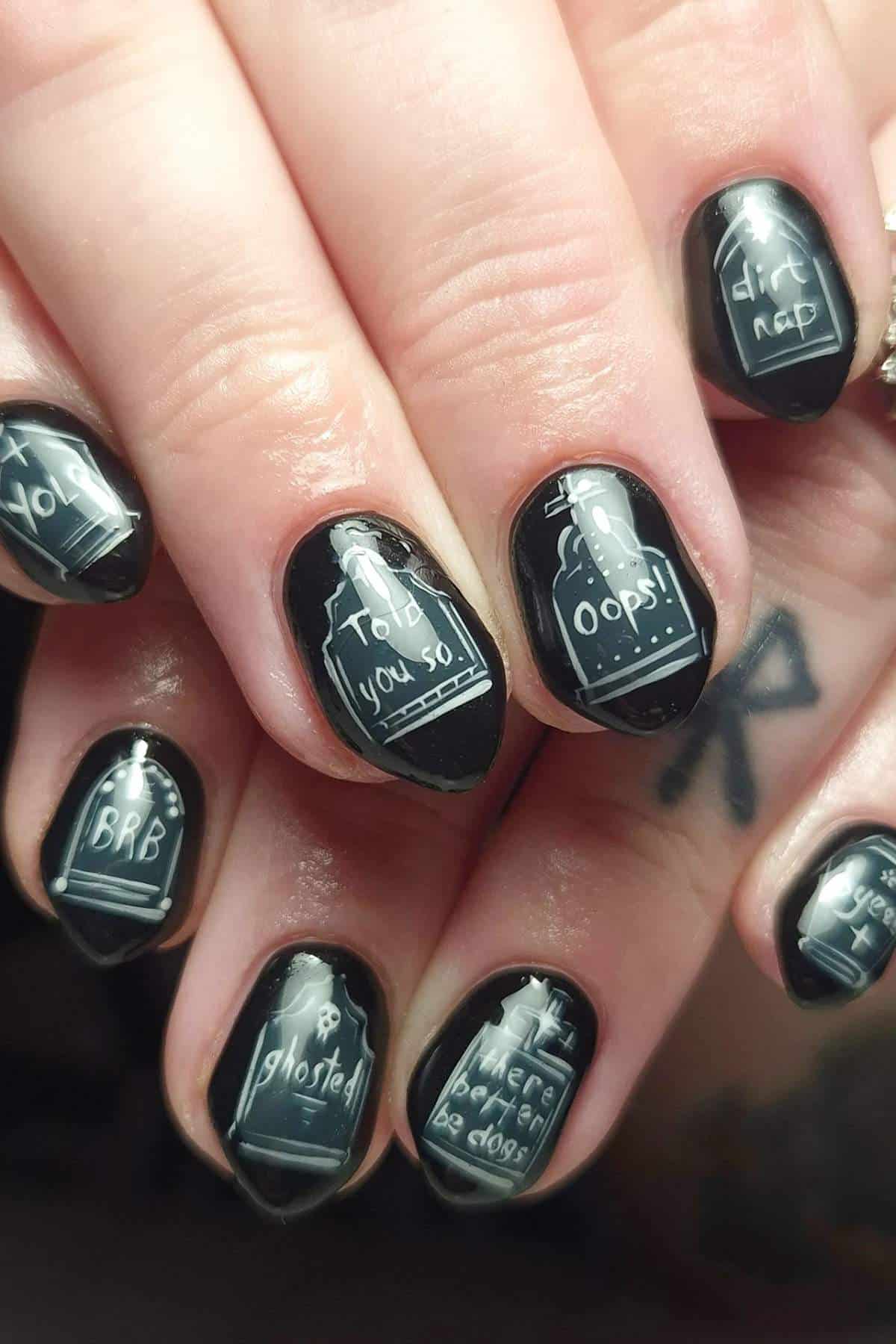 Close-up of black fingernails with white tombstone designs and phrases like "BRB," "Oops!," "I told you so!," and "Ghosted" written on them.