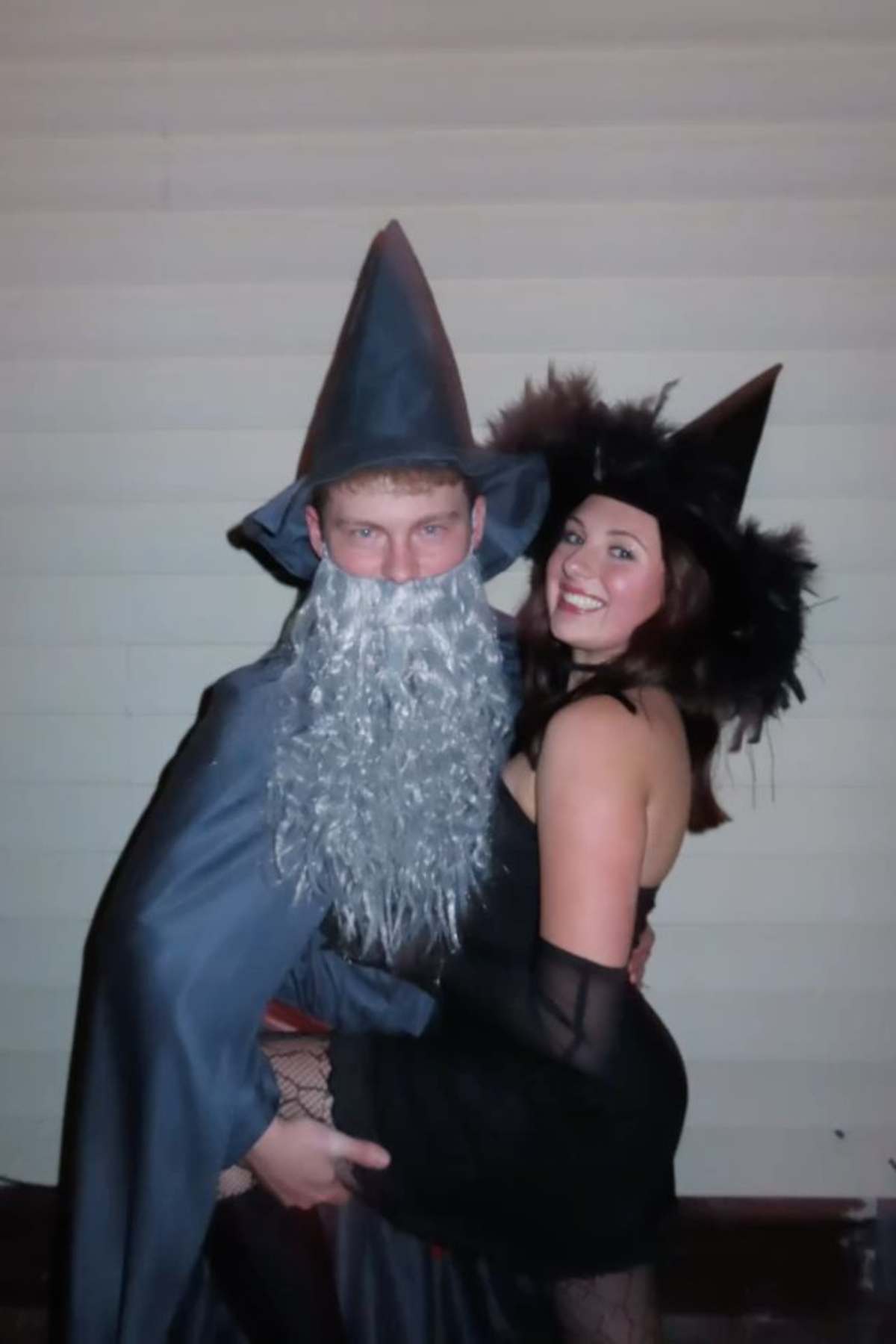 Two people in wizard and witch costumes pose together indoors, one wearing a gray pointed hat and beard, the other in a black hat and dress, both looking at the camera.
