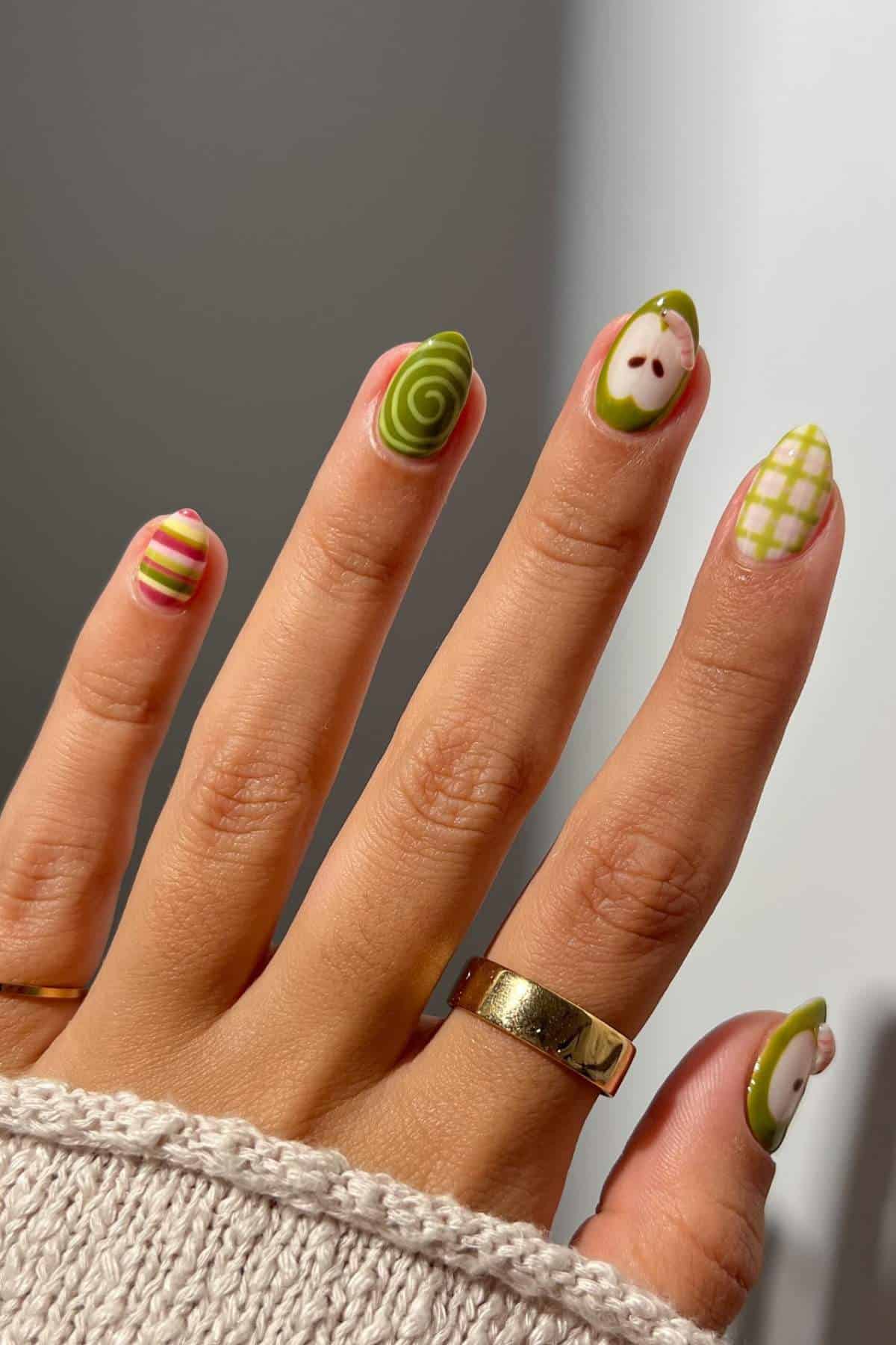 A hand with five fingernails painted in green, yellow, white, and pink designs, including stripes, swirls, checkerboard, and an abstract face.
