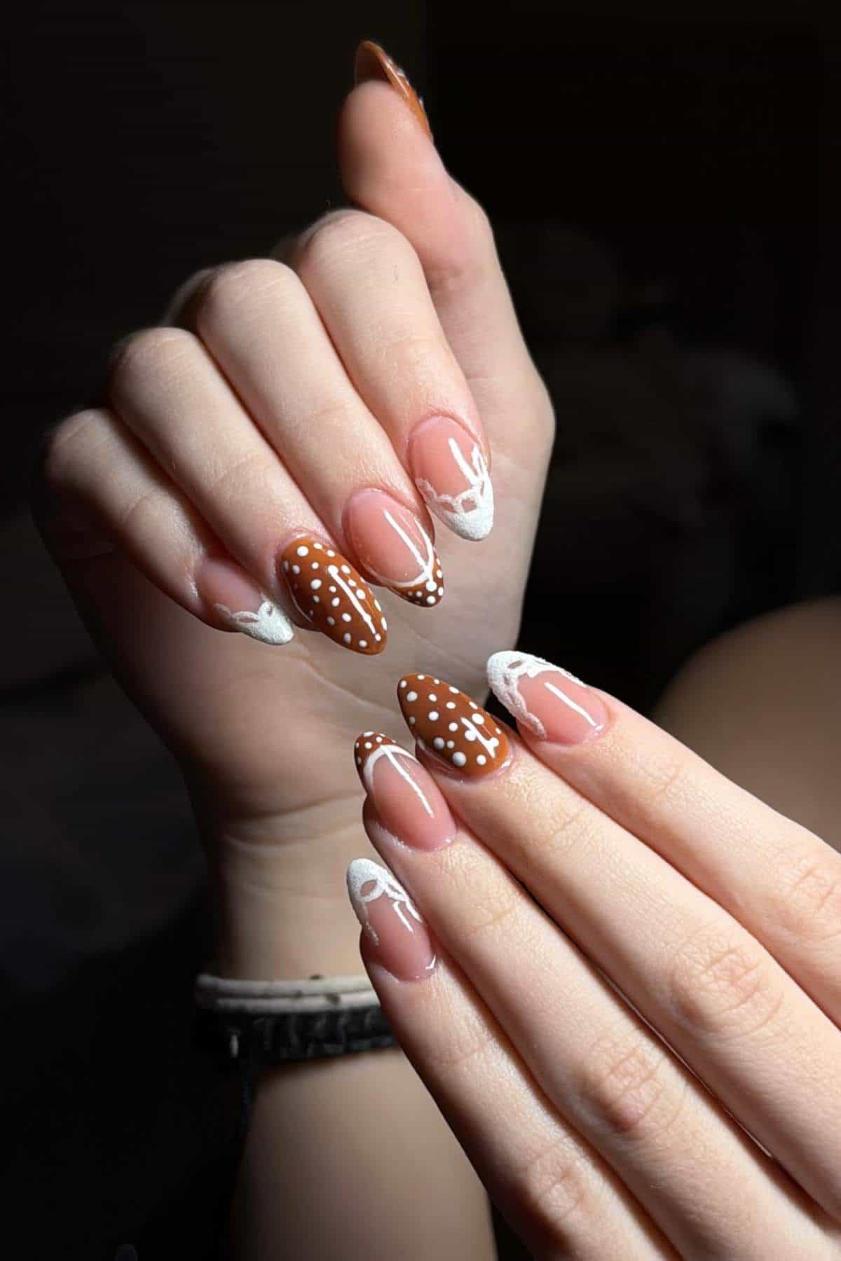 Hands with long, almond-shaped nails featuring brown and white deer-inspired nail art, including polka dots and white tips.