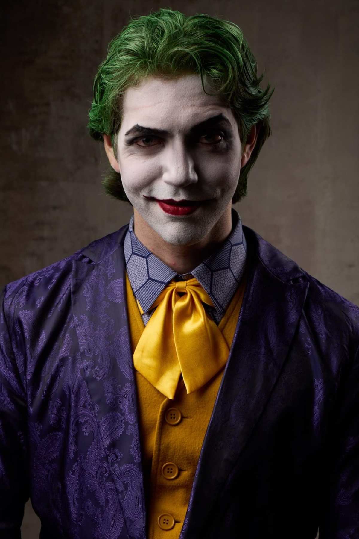 Person dressed as the Joker with green hair, white face paint, red lips, and purple and yellow suit, standing against a plain background.