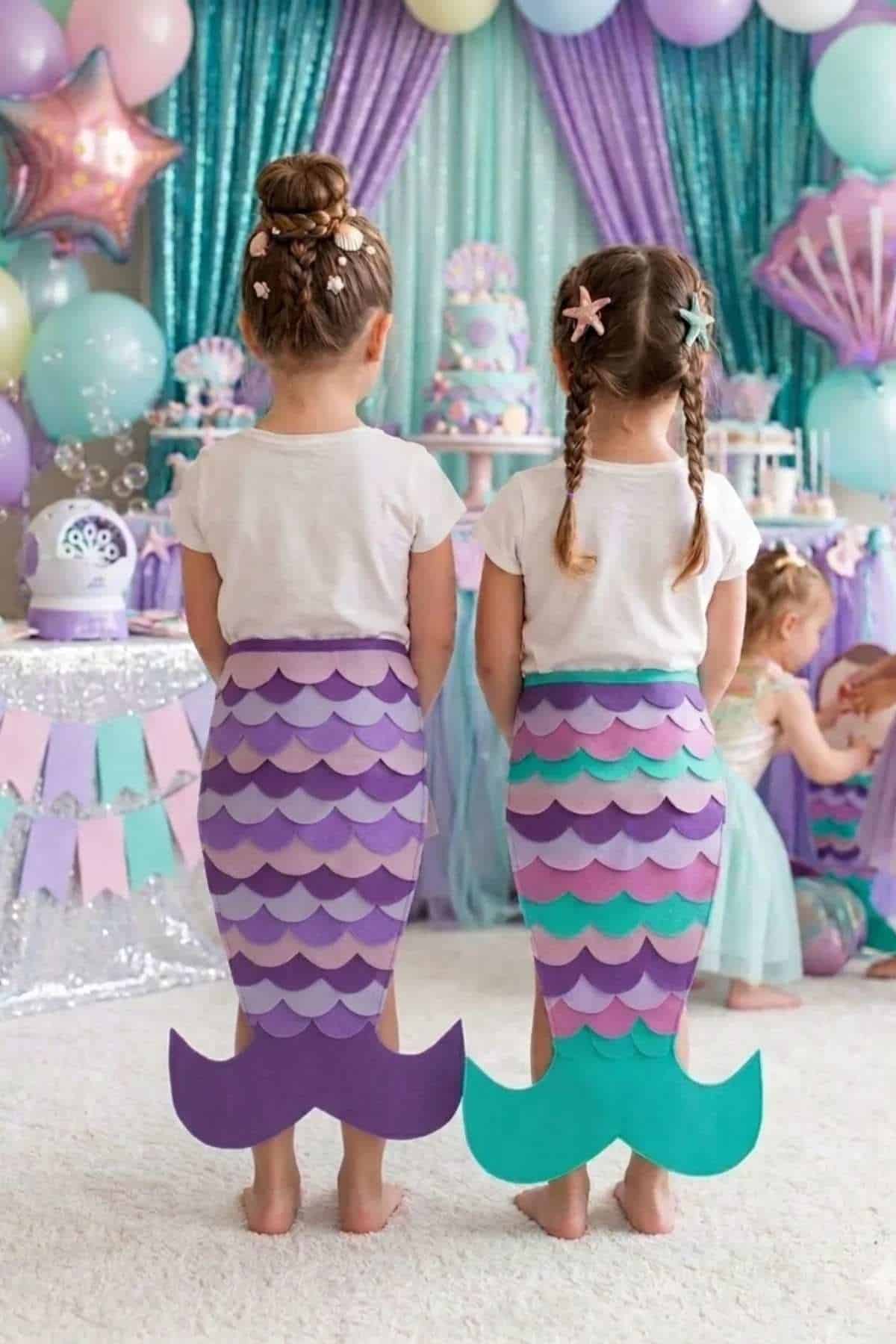 Two young children with braided hair wear mermaid tail costumes at a pastel-themed party with balloons, streamers, and other children in the background.