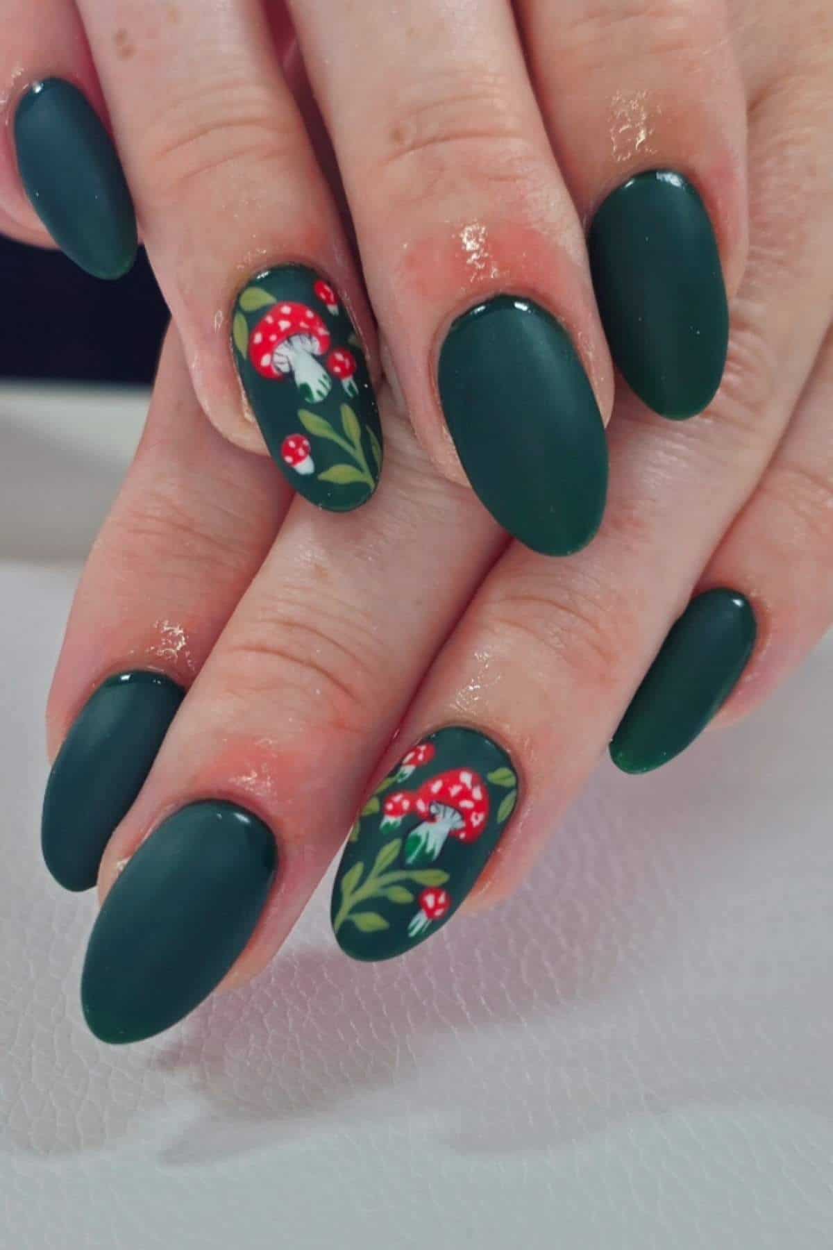 Hands with matte dark green oval nails; ring fingernails feature small red and white mushroom designs with green leaves.