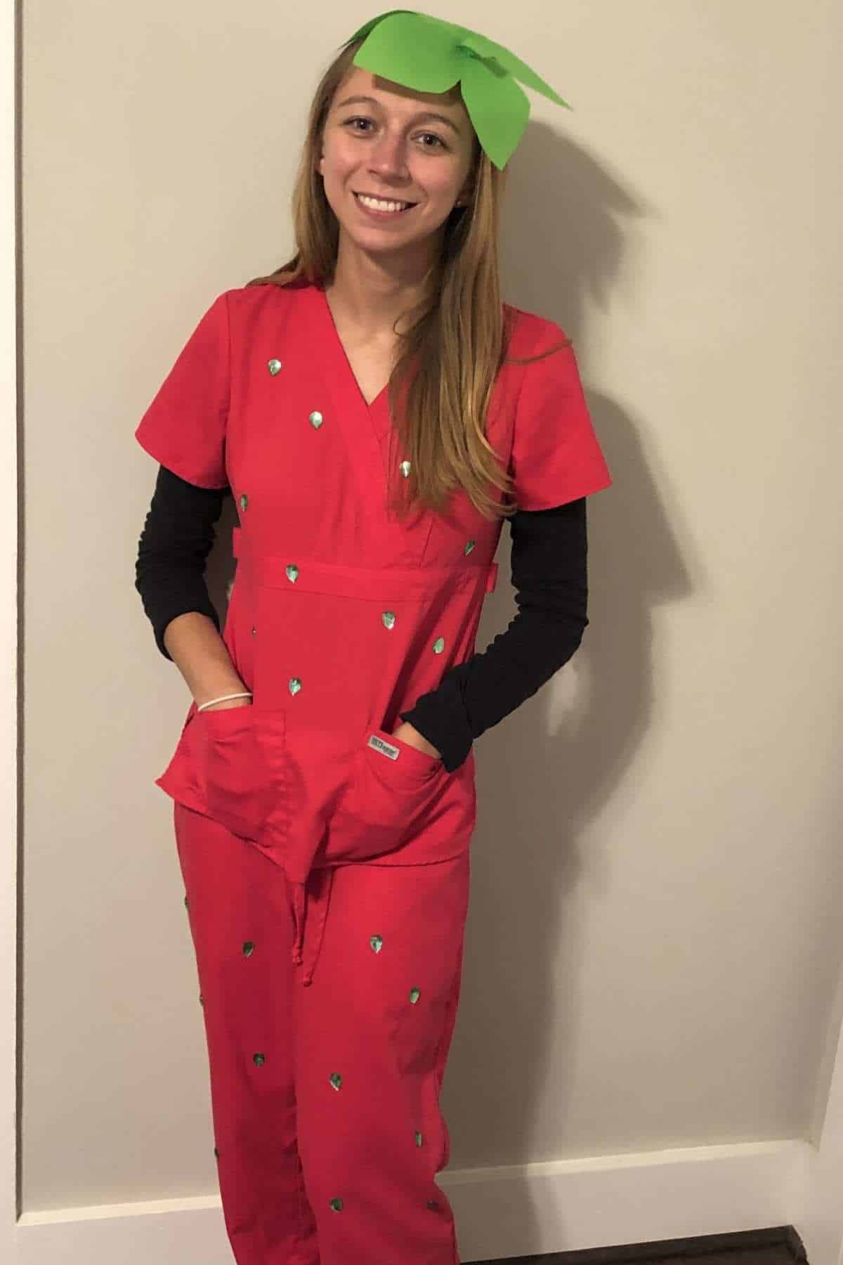 Person wearing a red jumpsuit with green spots and a green paper leaf headpiece, standing against a plain wall, resembling a strawberry costume.