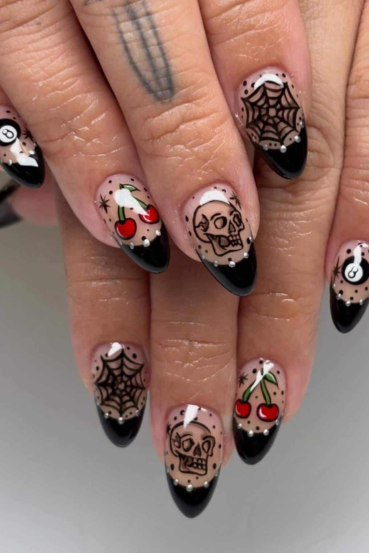 Close-up of hands with almond-shaped nails featuring black French tips and designs of skulls, cherries, spiderwebs, and 8-balls on a nude base.