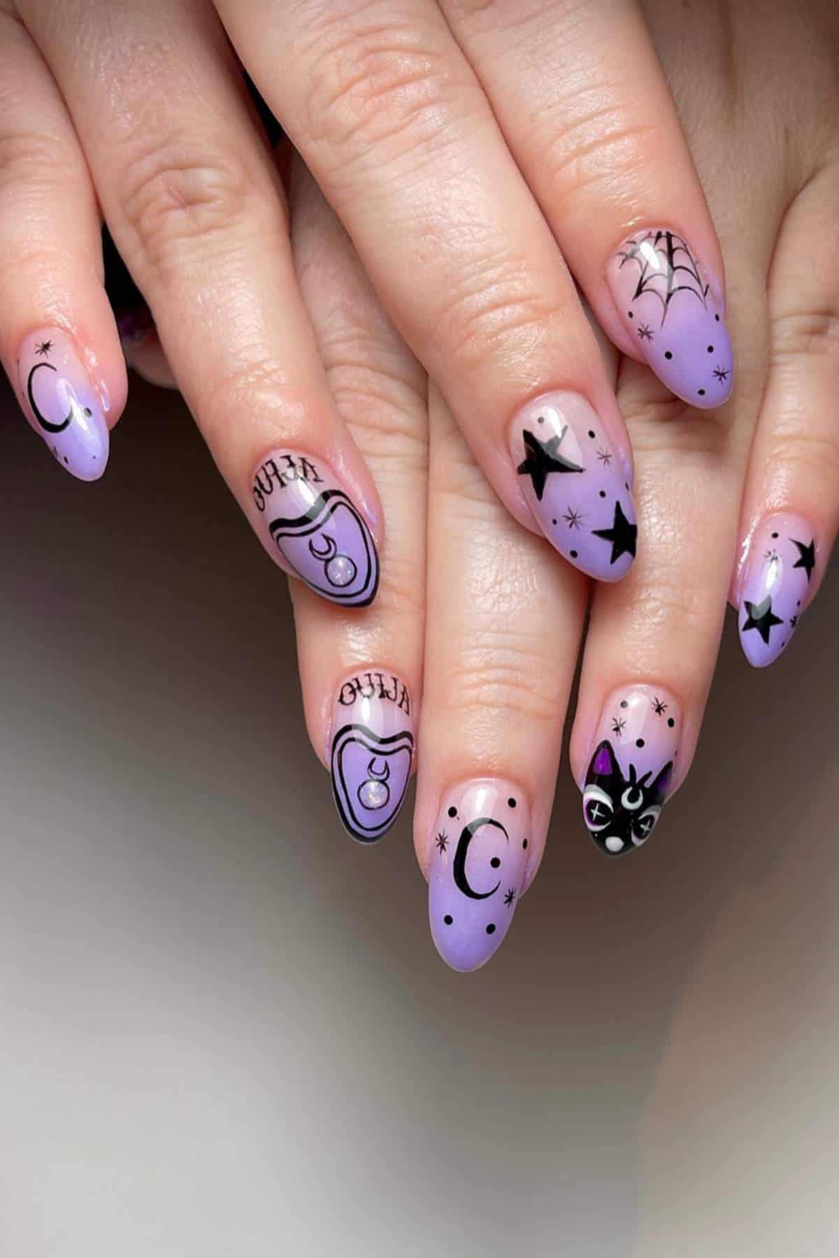 Hands with long, almond-shaped nails painted purple and decorated with black stars, crescent moons, a cat face, spider web, and the words "weird" and "creep.