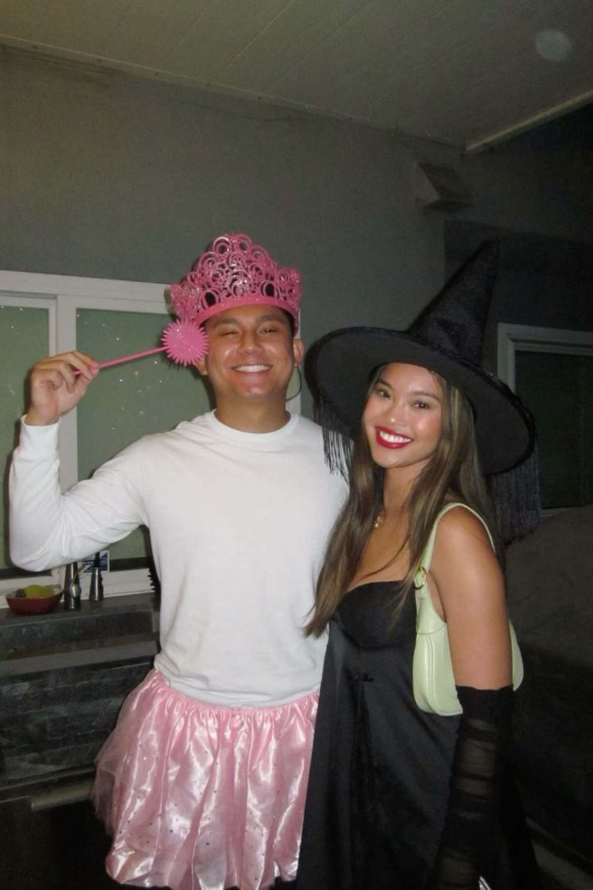 Two people in costumes: one wears a pink tiara, wand, and tutu; the other wears a black witch hat and dress. They are smiling and standing indoors.