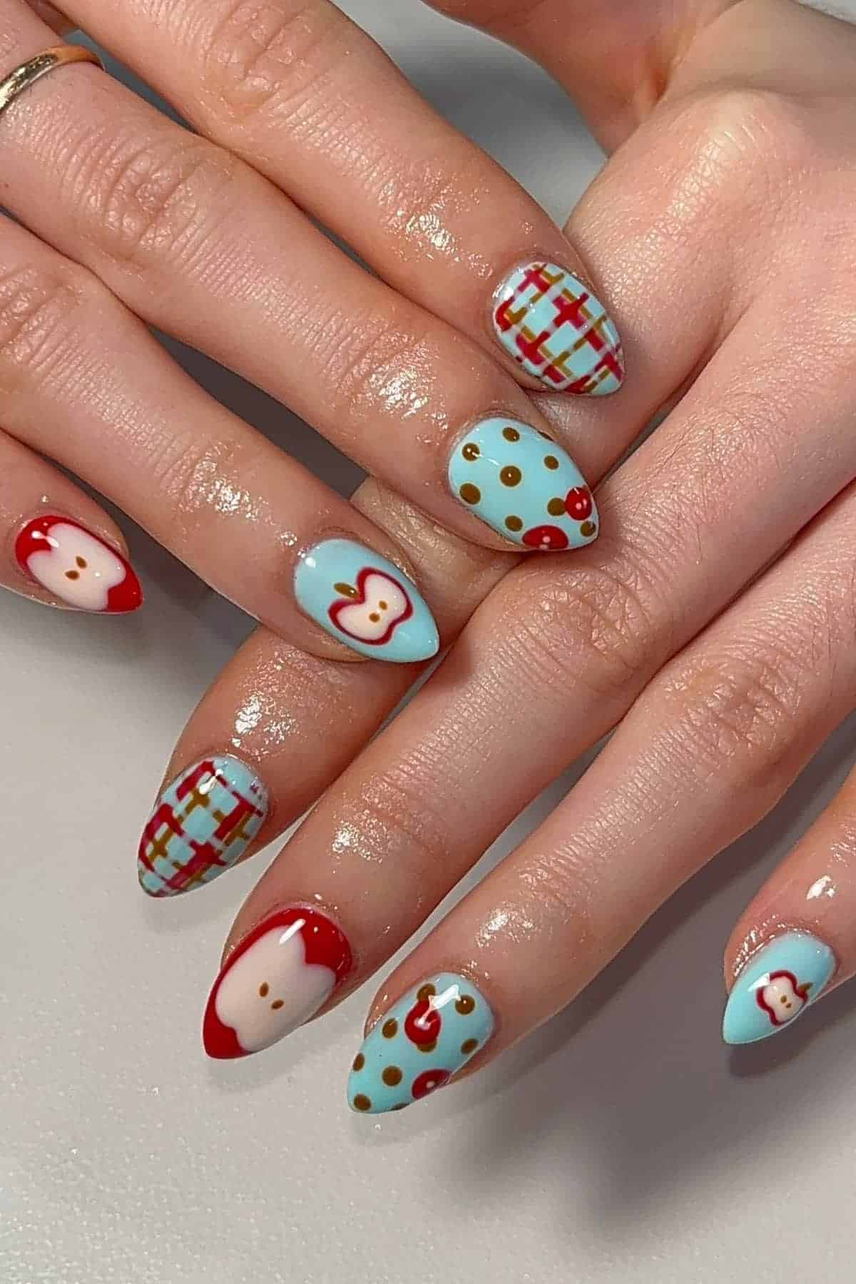 A hand with almond-shaped nails featuring blue, red, and beige nail art with polka dots, plaid patterns, and cartoon faces.