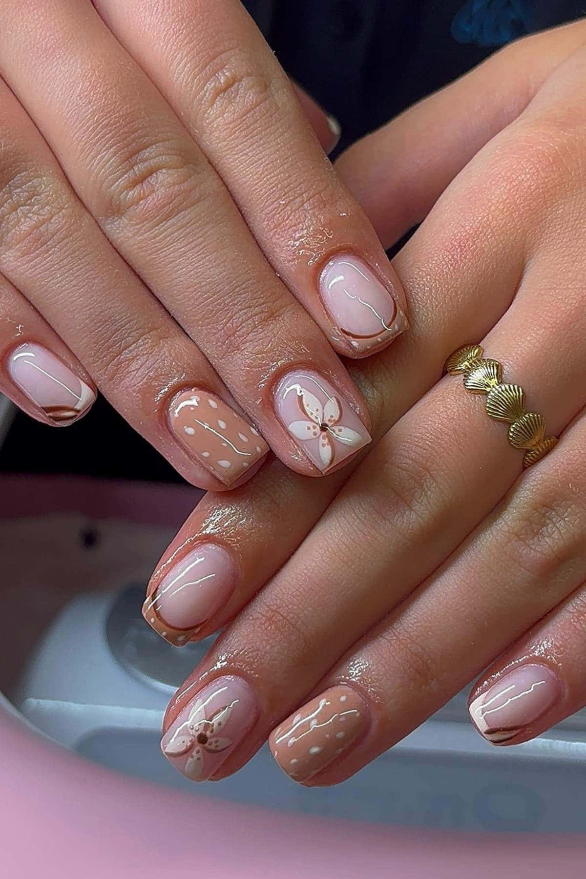 Hands with short, square-shaped nails painted with nude polish, featuring white dotted lines, floral designs, and metallic gold accents. Person is wearing a gold ring on one finger.