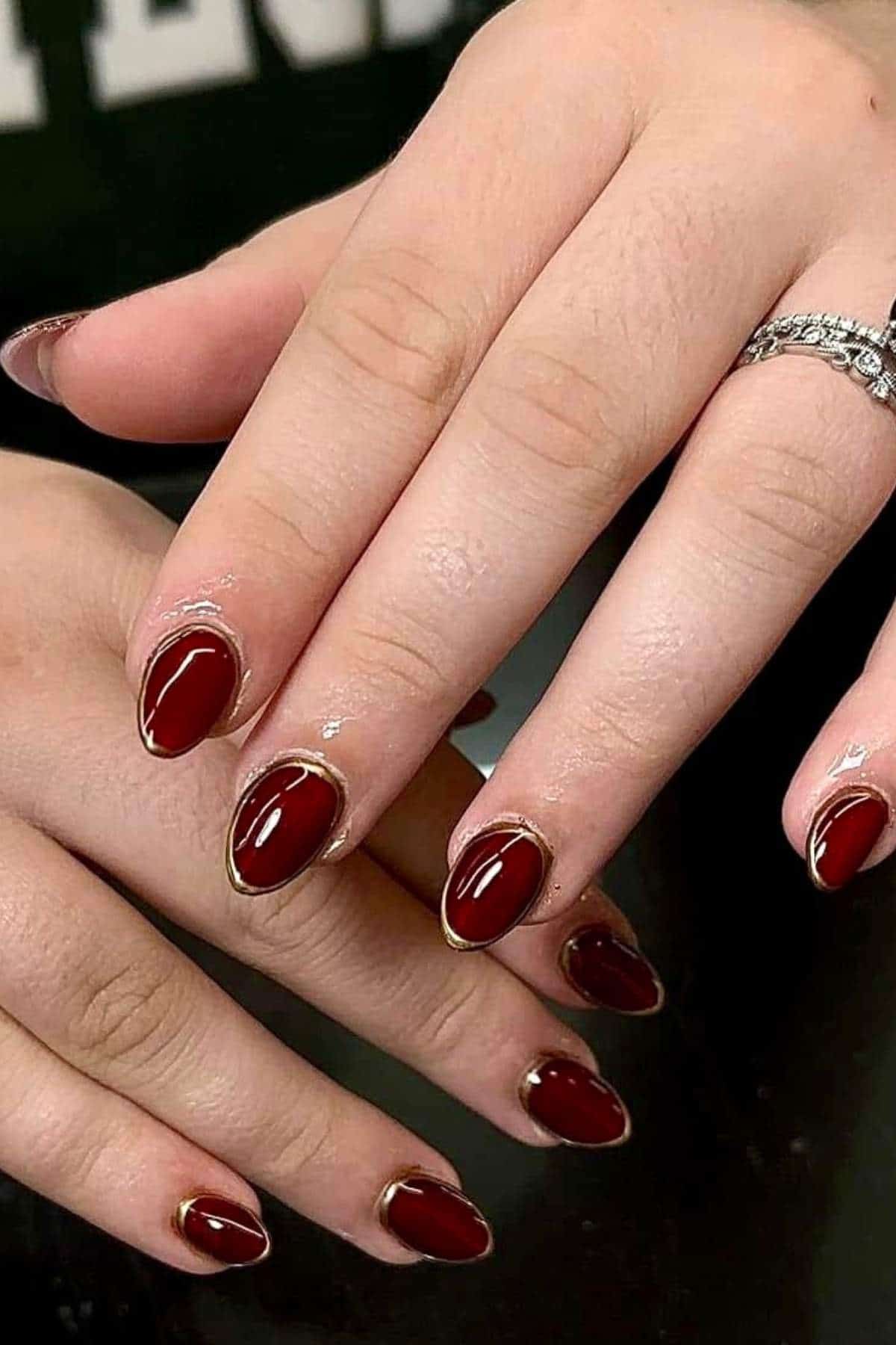 A pair of hands with almond-shaped nails painted dark red with a thin gold outline near the cuticle. The person is wearing a silver ring on one finger.