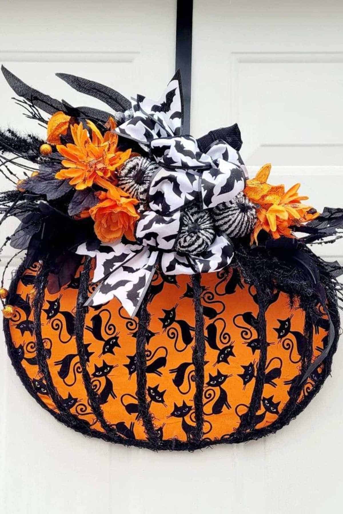 Orange pumpkin wreath with black cat silhouettes, black trim, orange flowers, and black-and-white patterned bows; hangs on a white door.