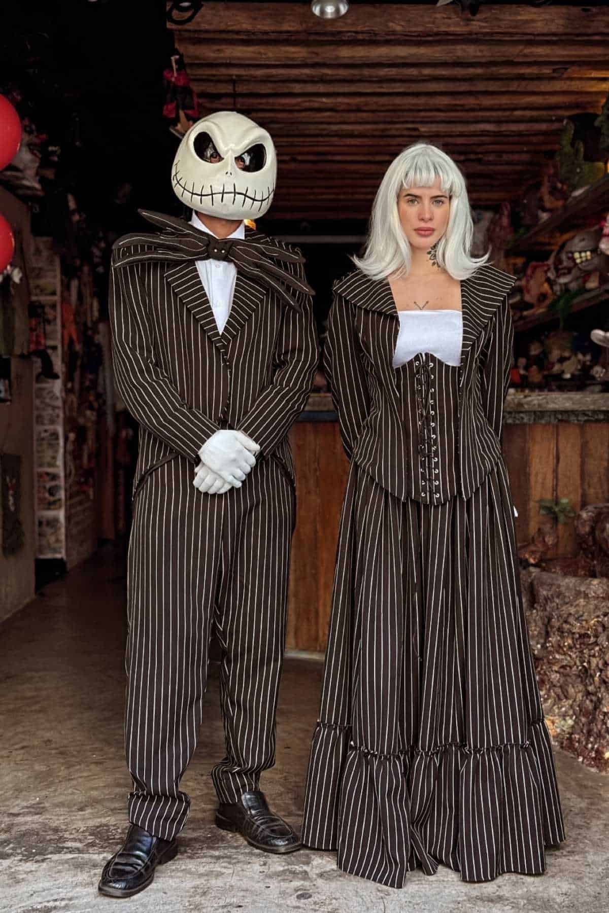 Two people in black and white pinstripe costumes, one wearing a Jack Skellington mask and gloves, the other with long white hair, standing side by side indoors.