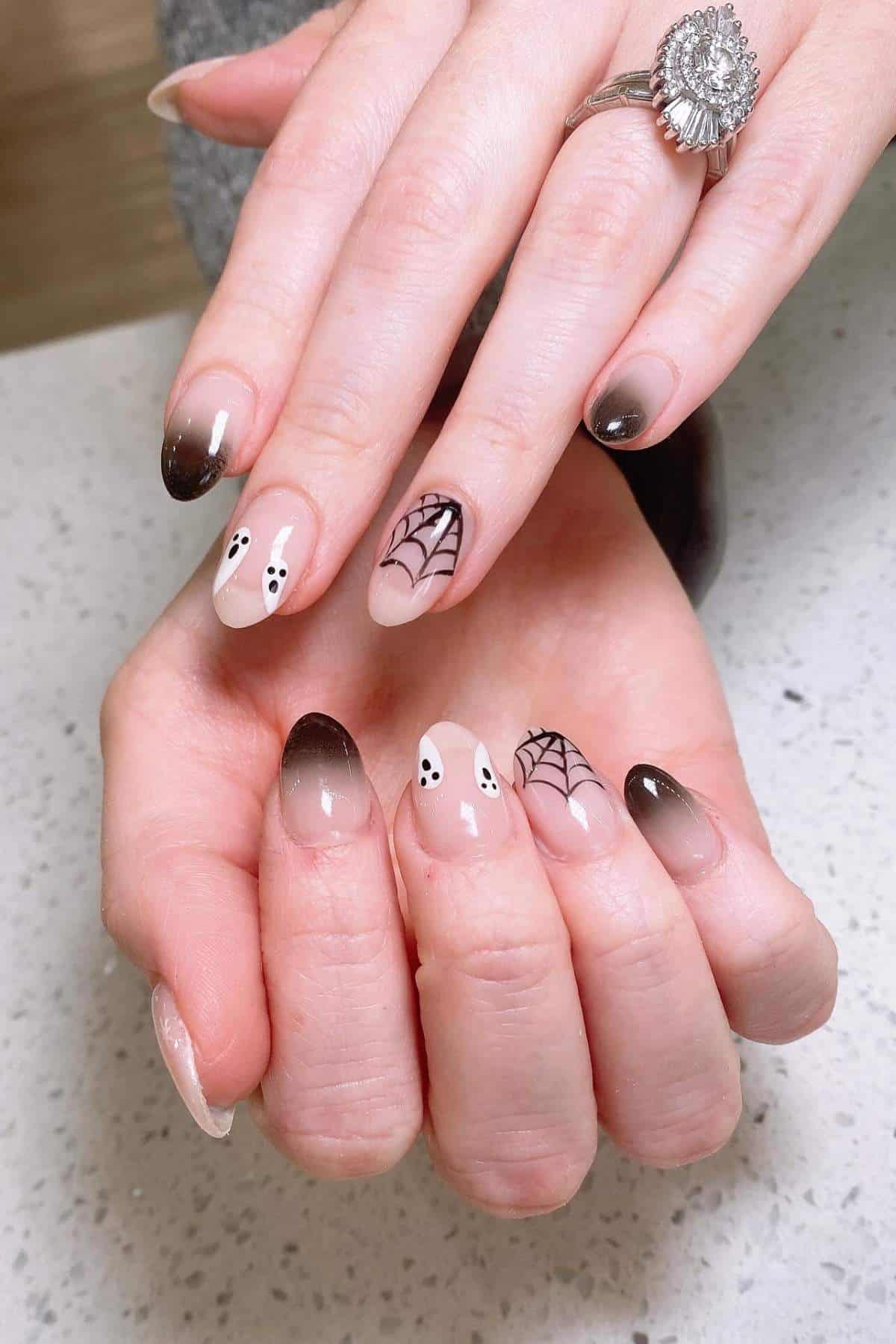 Hands with almond-shaped nails featuring brown ombré tips, ghost and spiderweb nail art, resting on a light speckled surface.