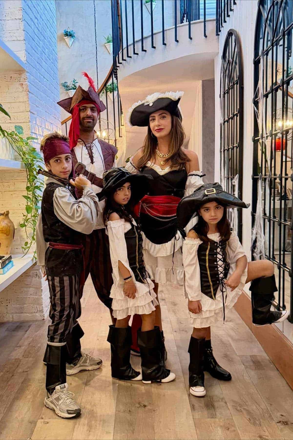 A family of five poses indoors wearing matching pirate costumes, including hats, boots, and striped or frilled outfits, in a modern, well-lit home with plants and arched windows.
