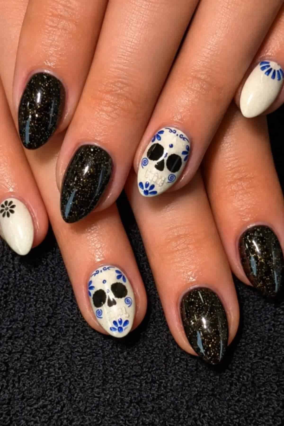 A hand with black glitter, white, and skull-themed nail art featuring blue floral accents and small black designs on the nails.