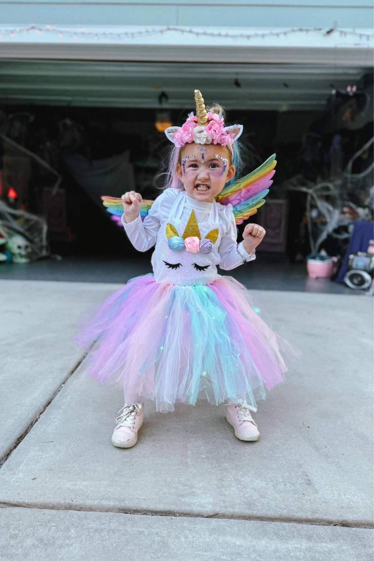 A young child in a unicorn costume with rainbow wings, a horn headband, and a tutu stands on a driveway in front of a decorated garage, posing enthusiastically.