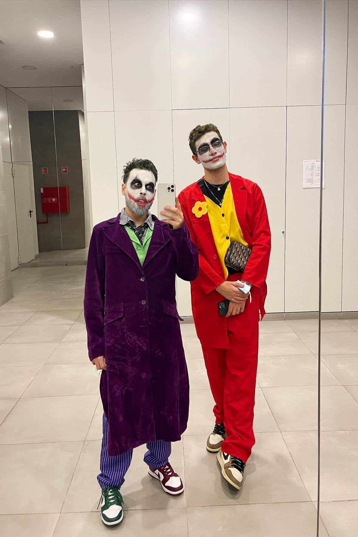 Two people dressed as the Joker take a mirror selfie in a brightly lit restroom, wearing colorful suits and face paint.
