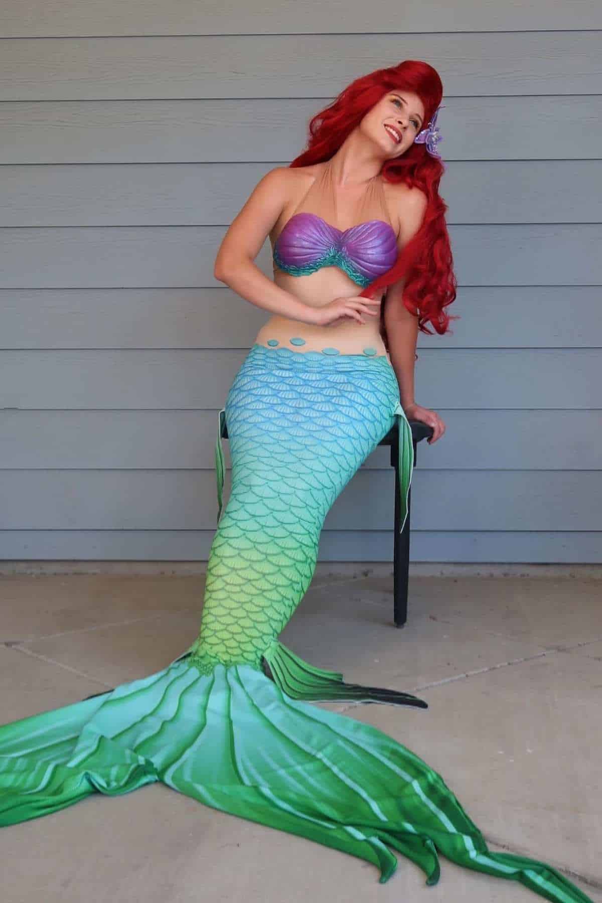 Person dressed as a mermaid with a red wig, purple seashell top, and green tail, sitting on a chair against a gray wood-paneled wall.