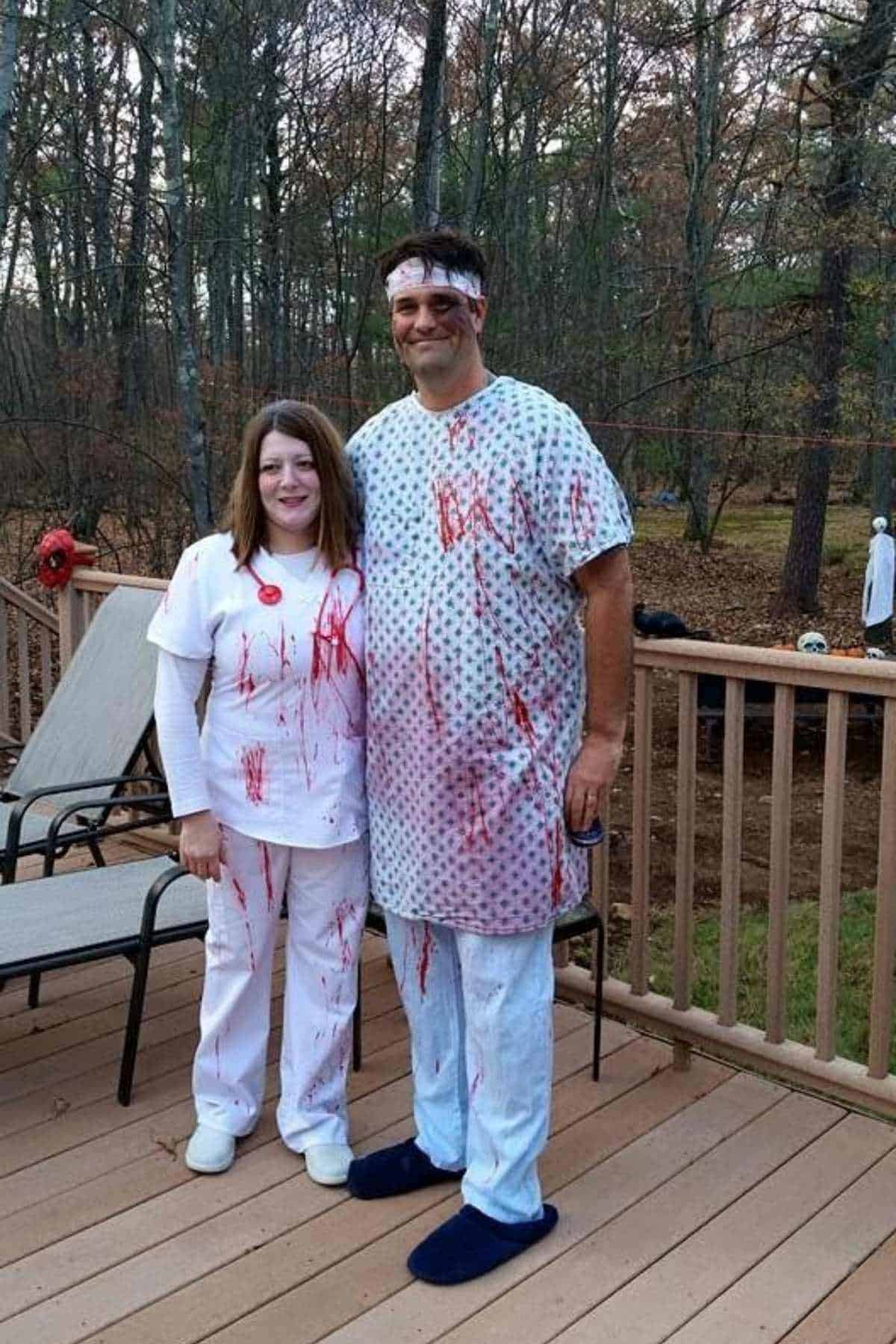 Two adults in medical-themed costumes splattered with fake blood stand on a wooden deck with trees in the background.