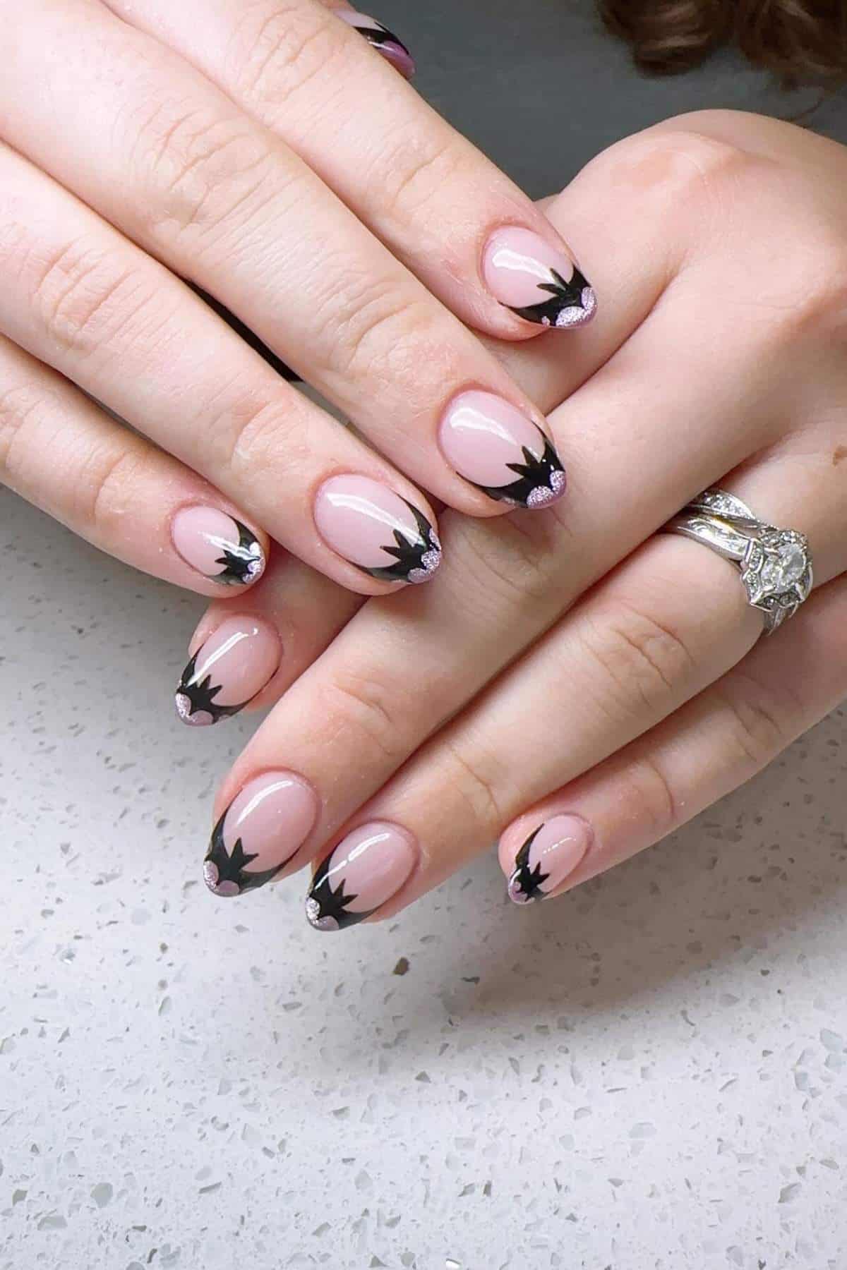 Hands with short, almond-shaped nails featuring a pink base and black floral-like tips, resting on a white speckled surface. A silver ring is worn on one finger.