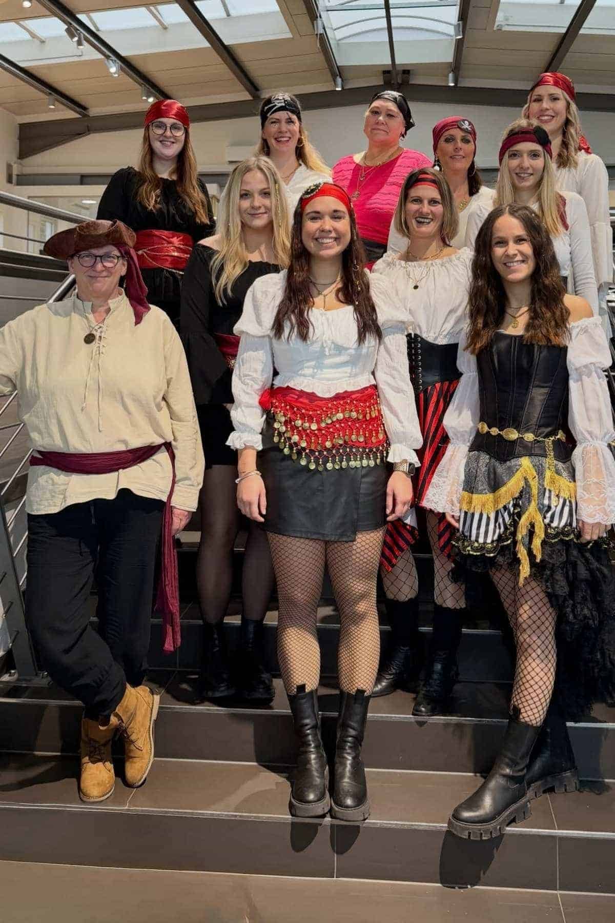 A group of twelve people in pirate-themed costumes pose together on a staircase, smiling at the camera indoors.