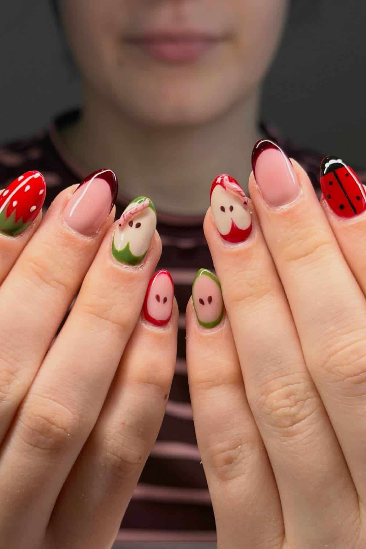 A person displays manicured nails with fruit-inspired and face designs in red, green, and nude tones. The background is out of focus.
