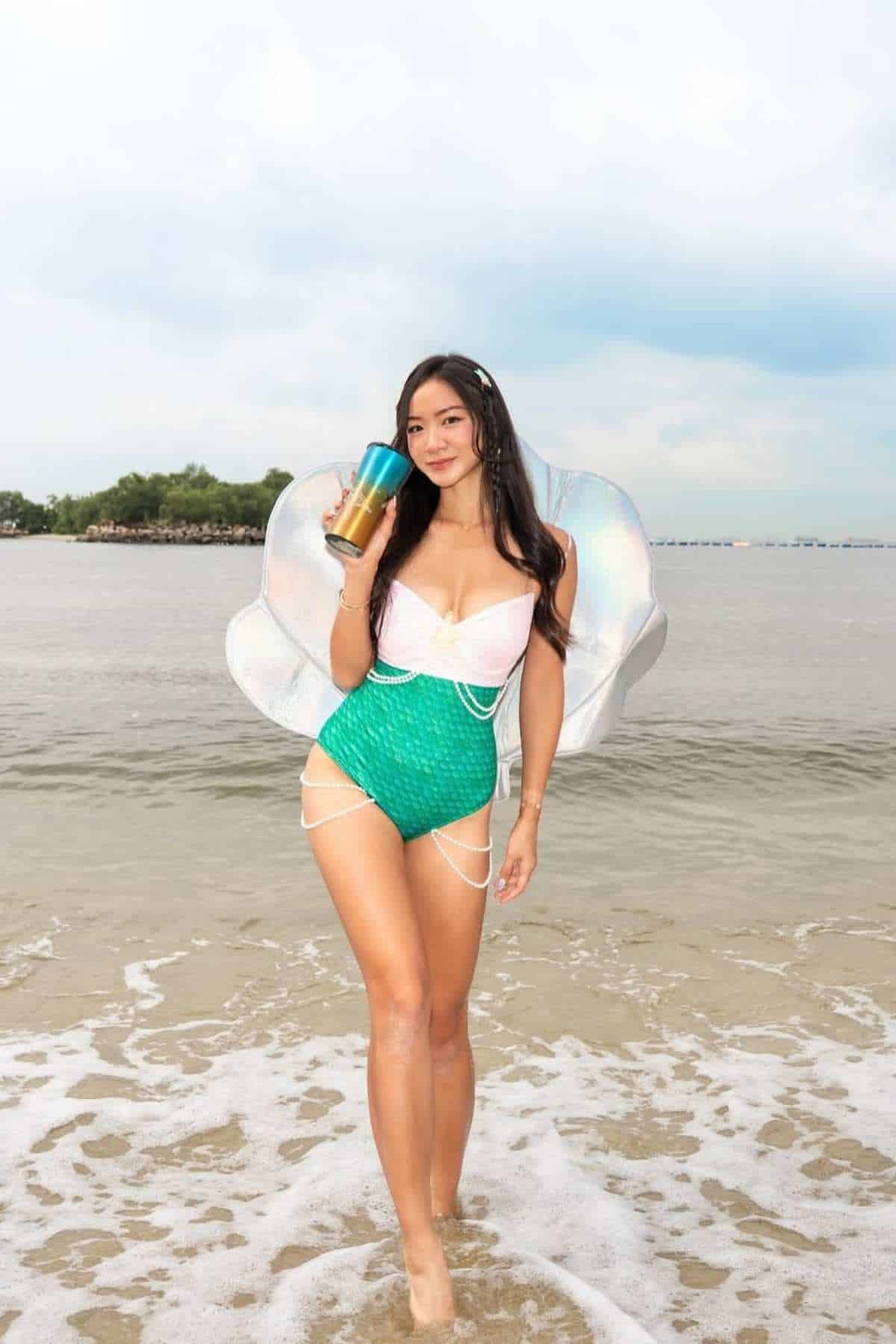 Woman in a mermaid-inspired outfit stands in shallow ocean water holding a drink, with a large shell prop behind her and shoreline visible in the background.