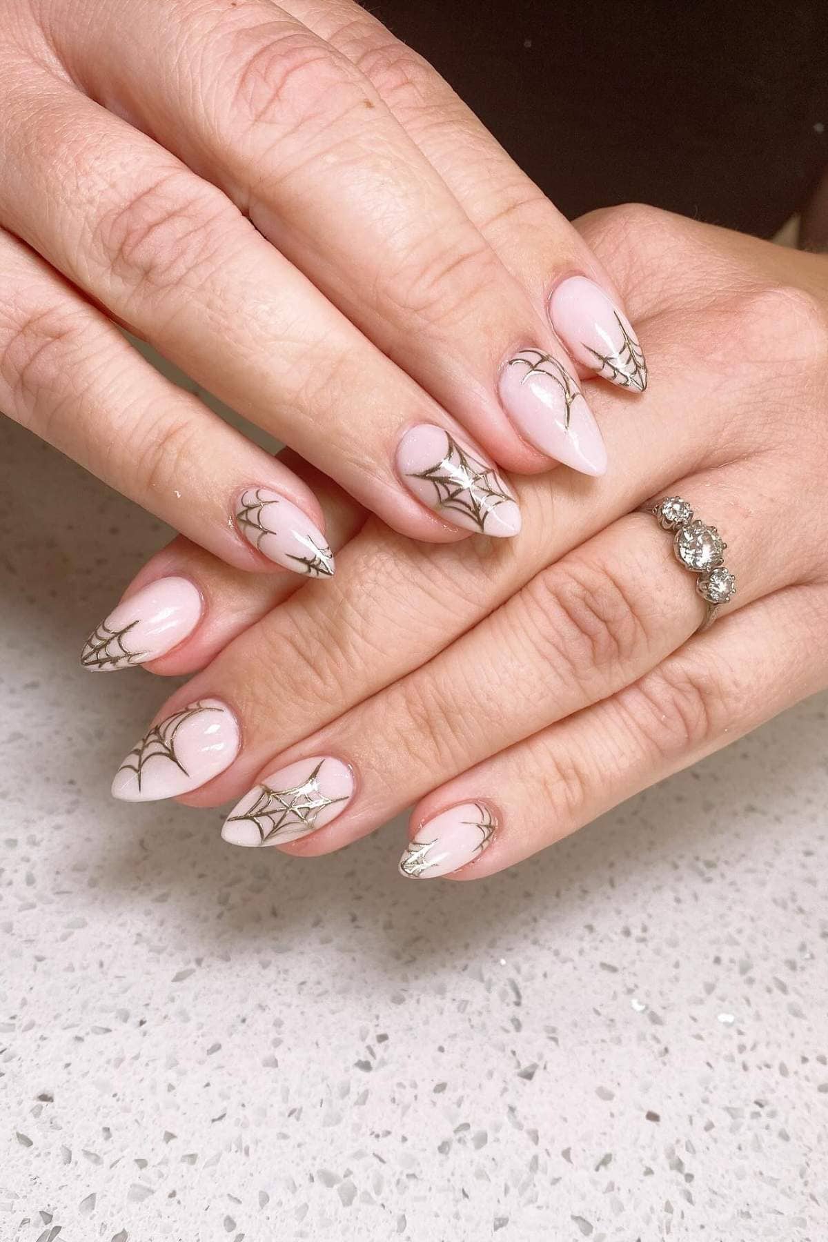 Hands with almond-shaped nails featuring pink polish and black spider web designs, resting on a speckled white surface. A ring with three stones is worn on one finger.