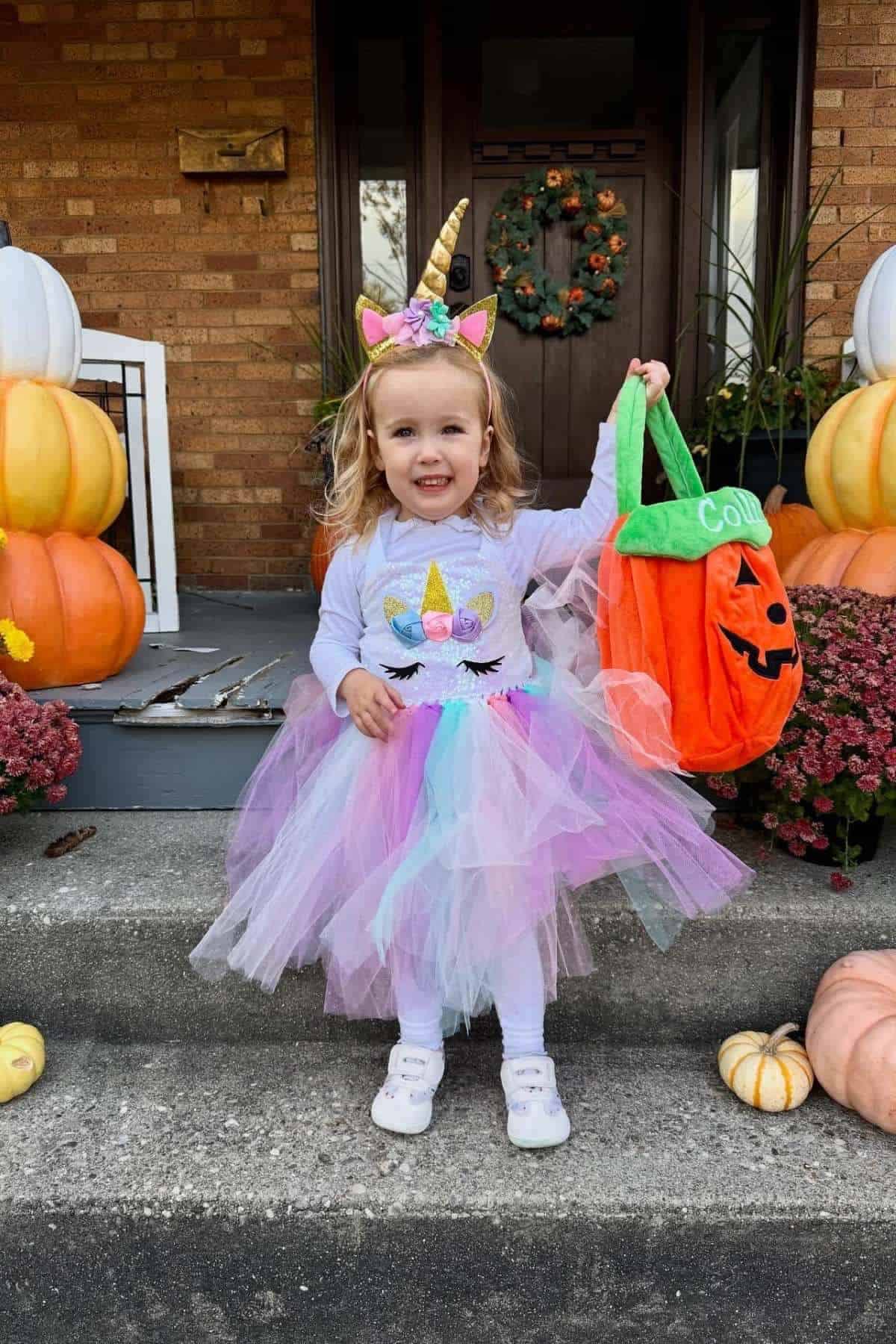Young child in a unicorn costume holds a pumpkin-shaped trick-or-treat bag on a decorated porch with pumpkins and flowers.
