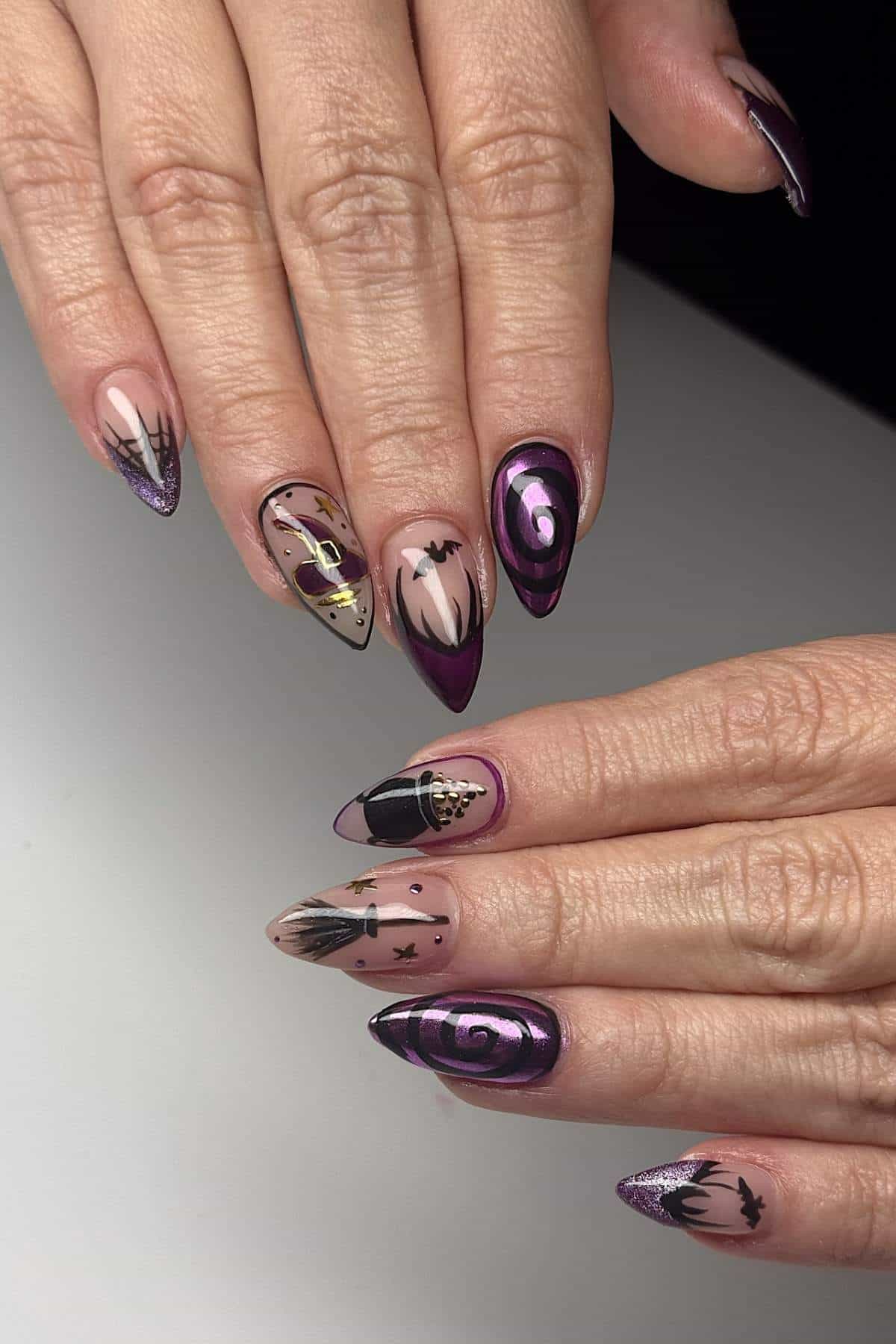 Hands with long, pointed nails featuring Halloween-themed designs: bats, spiderwebs, a witch, and swirling patterns in purple, black, and nude tones.