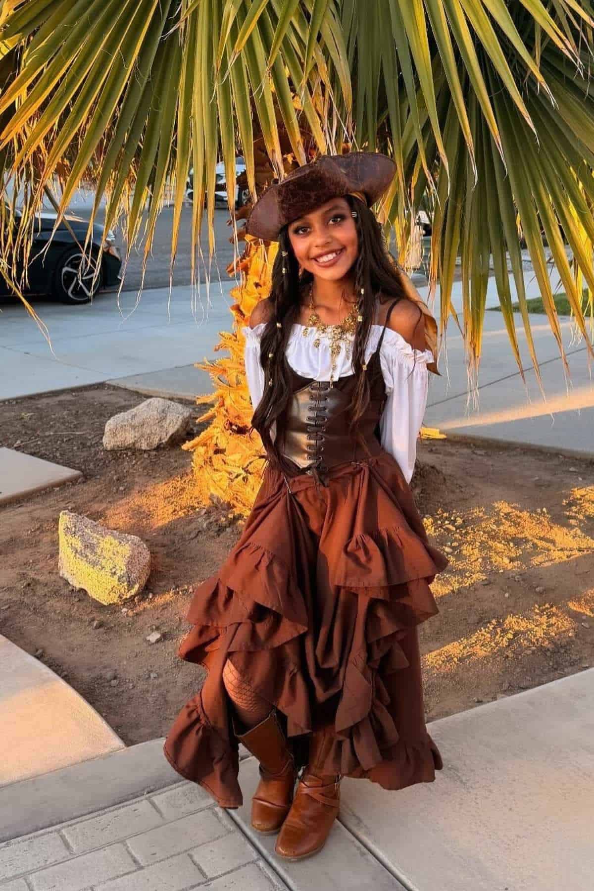 A woman stands outdoors in a pirate-style costume with a brown skirt, white blouse, hat, and boots, smiling in front of a palm tree.