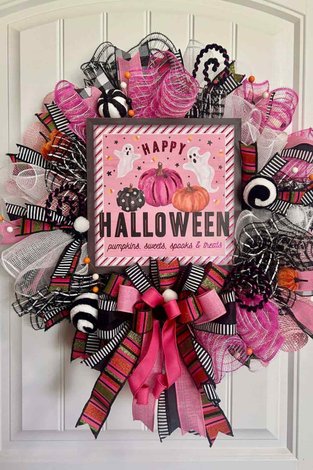 A pink and black Halloween wreath with a sign that says "Happy Halloween" and features ghosts, pumpkins, and candies, decorated with ribbons and mesh bows.
