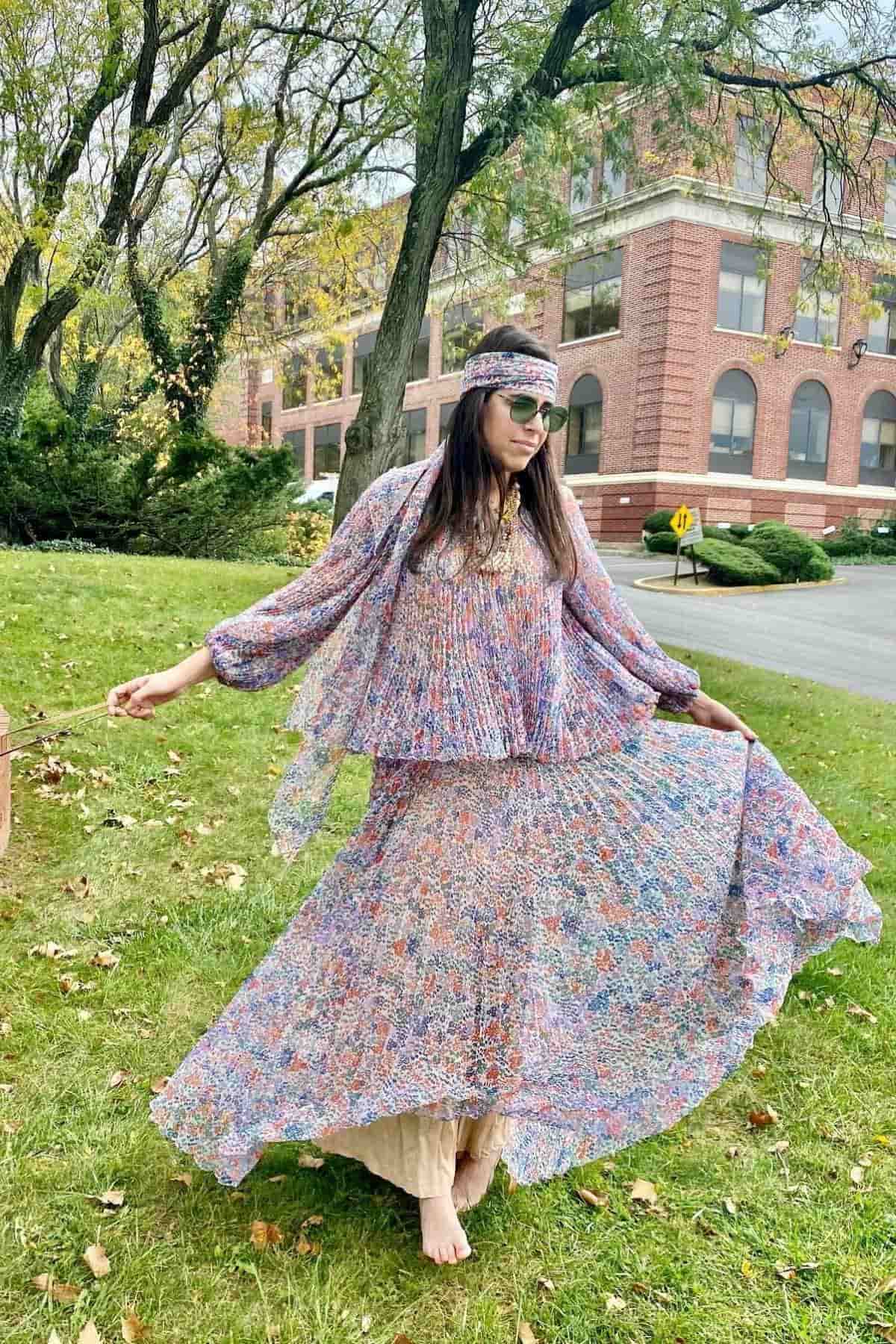 Woman in a colorful, flowy dress and headband stands barefoot on grass, holding her skirt out, with trees and a brick building in the background.