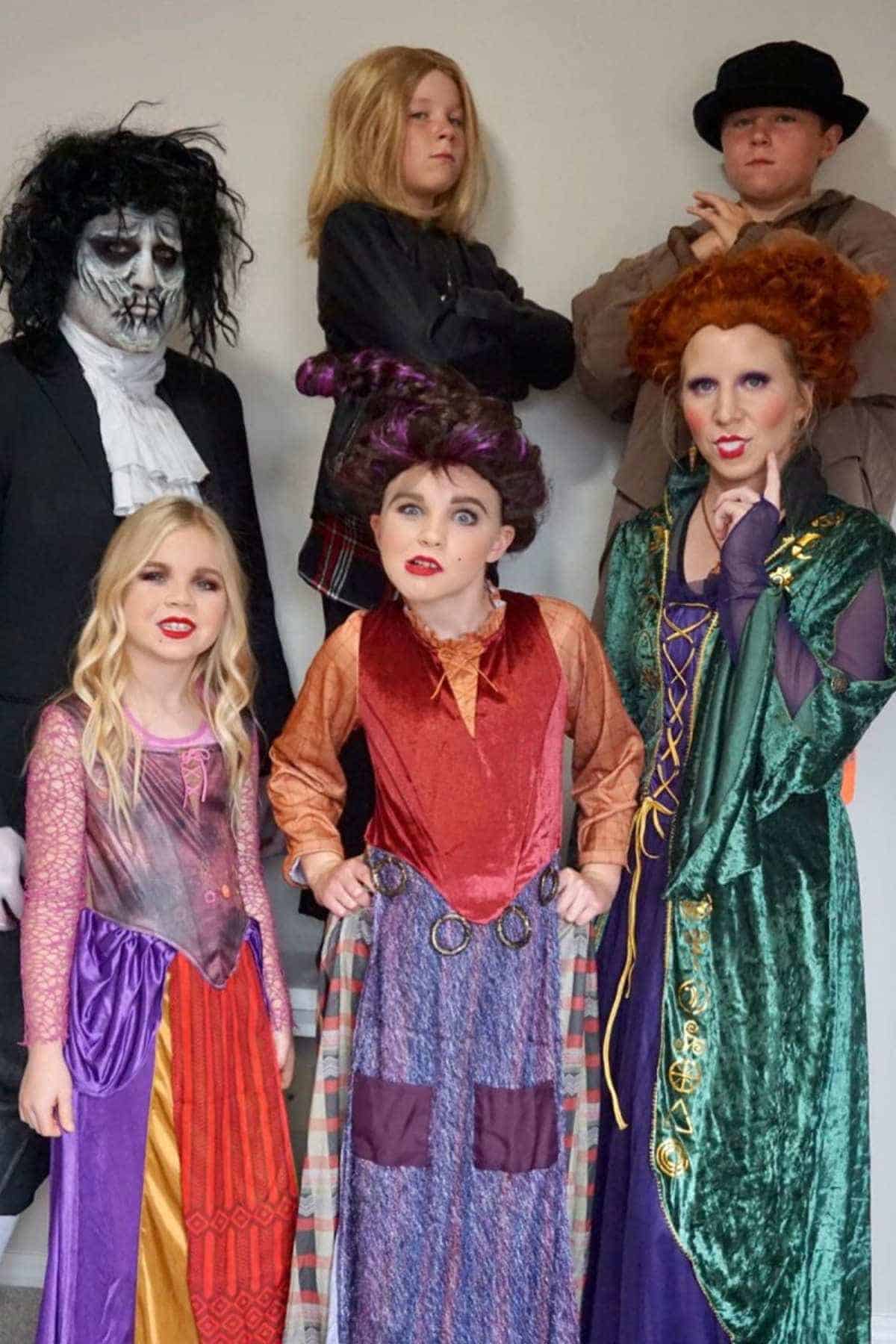 Six people in costume, four dressed as the Sanderson sisters from "Hocus Pocus," one in black with face makeup, and one in dark clothing and a hat, posing against a plain wall.