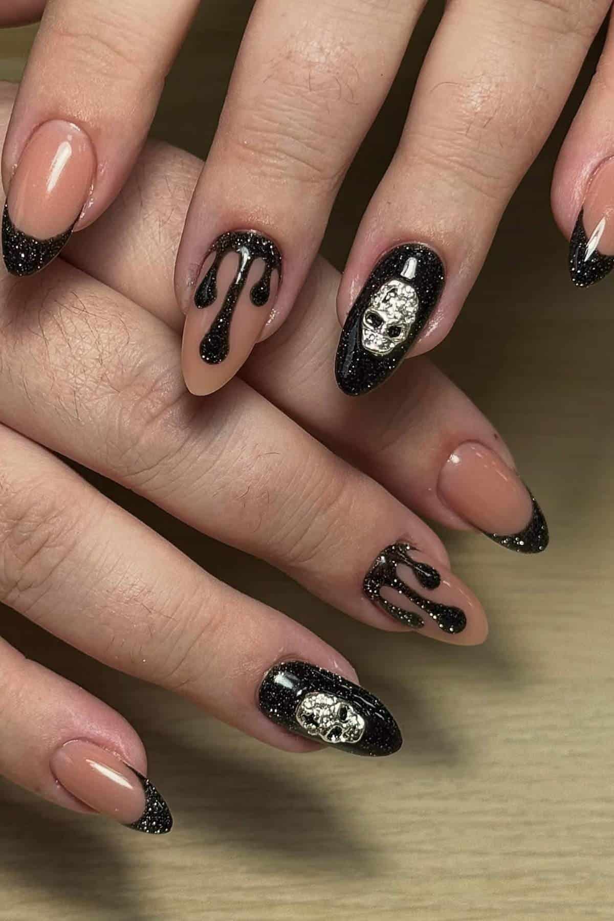 Hands with almond-shaped nails featuring nude polish, black glittery tips, dripping black designs, and metallic skull embellishments on some nails.