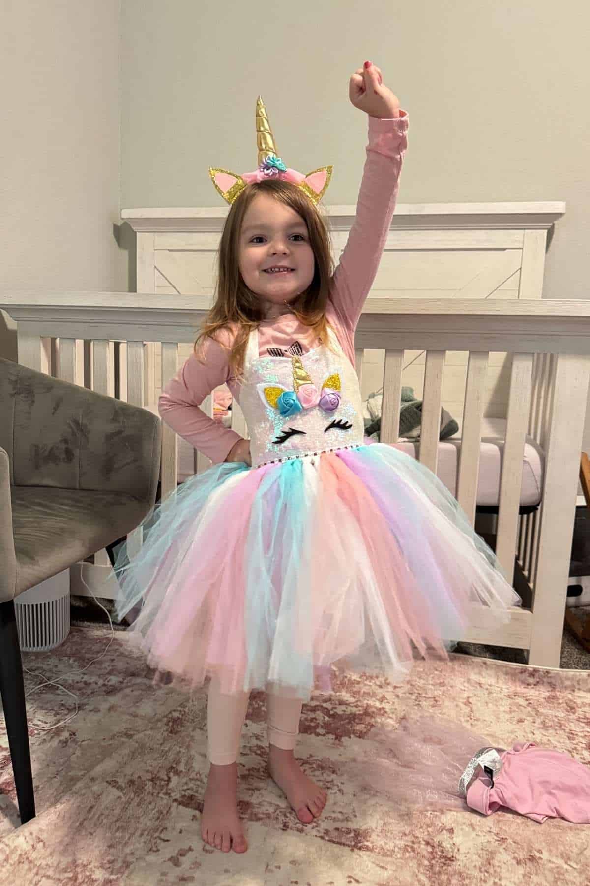 Young girl in a unicorn costume stands in a bedroom, smiling and raising one arm. A crib, chair, and pink rug are visible in the background.