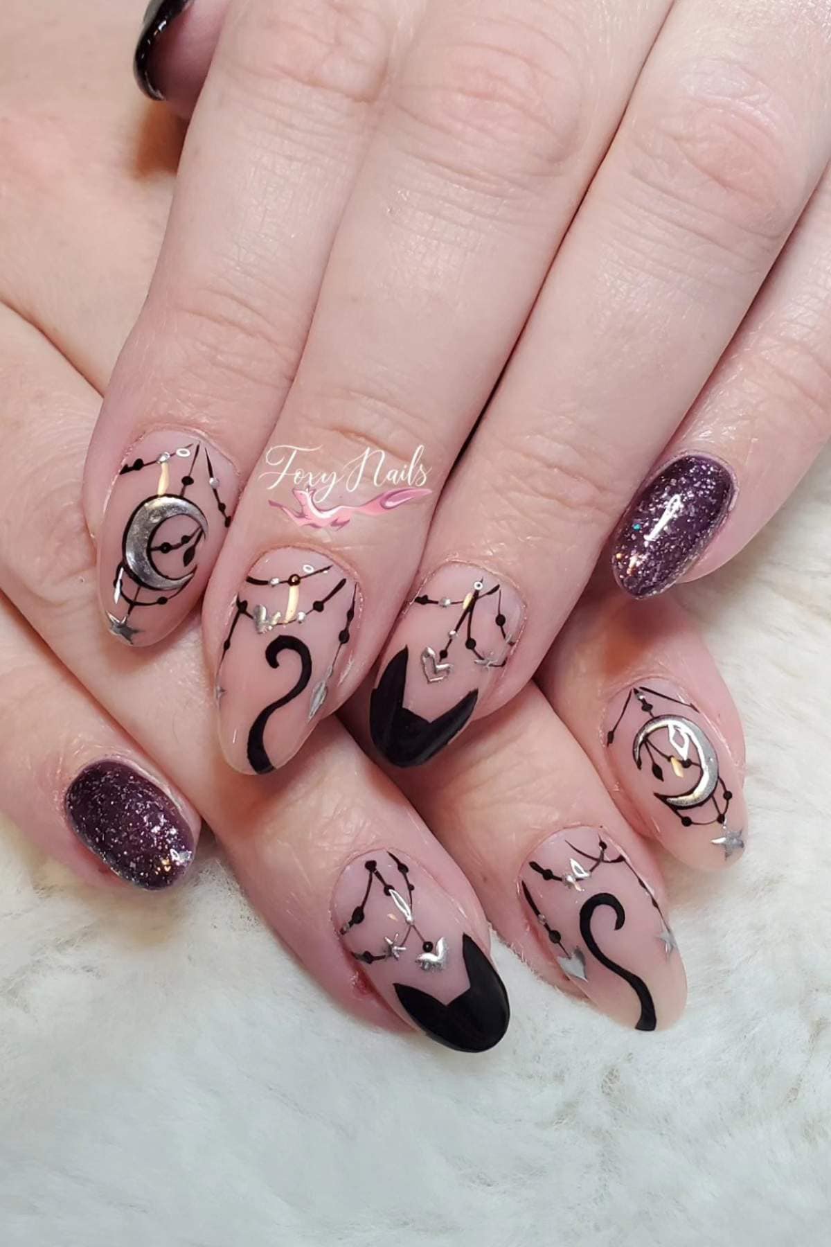 Hands with manicured nails featuring black abstract designs, silver decals, and purple glitter accent nails on a light background.