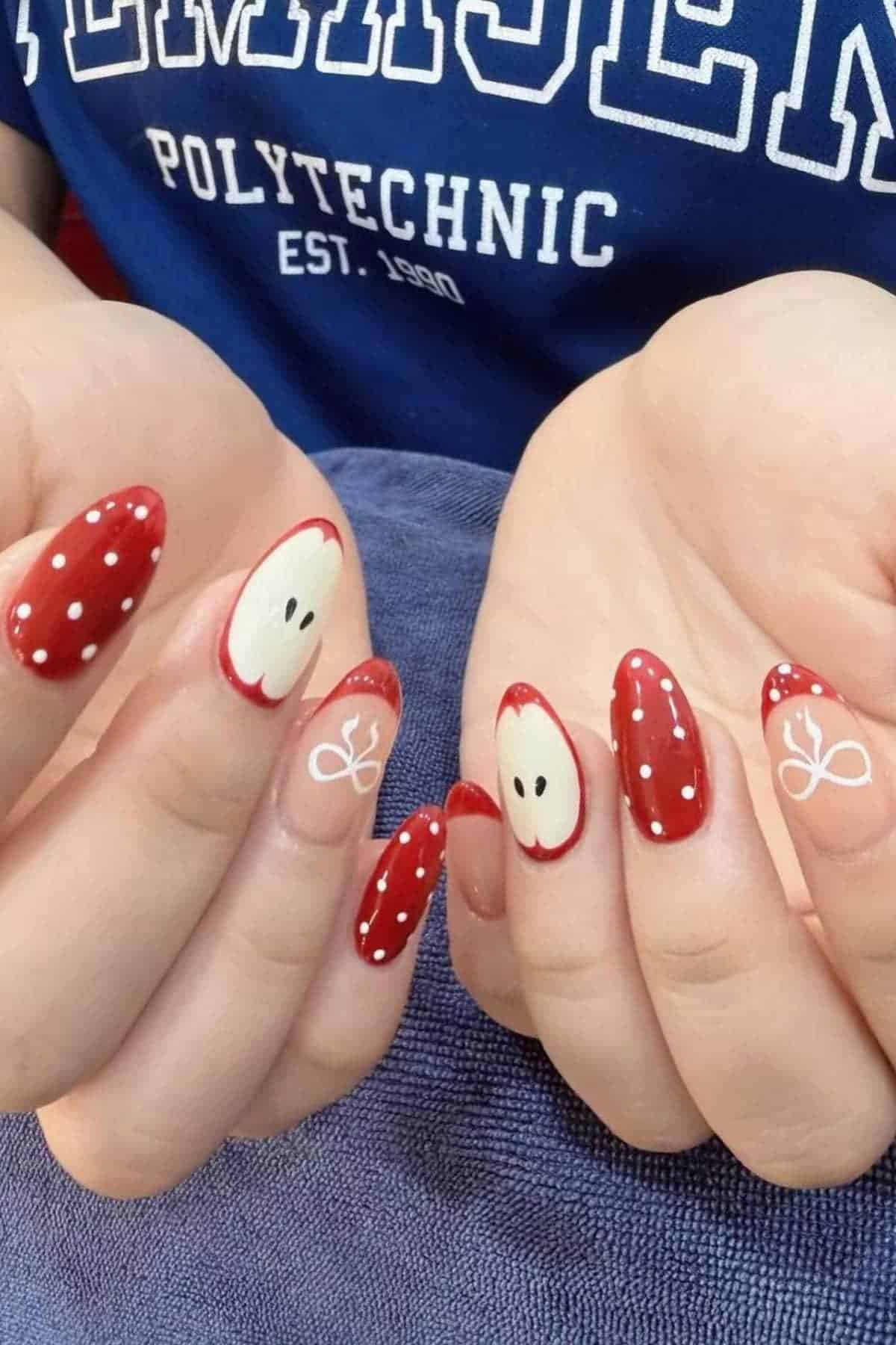 Hands with manicured nails featuring red polish with white polka dots, apple slice designs, and a white bow accent on a natural base. Person wears a blue shirt with white text.
