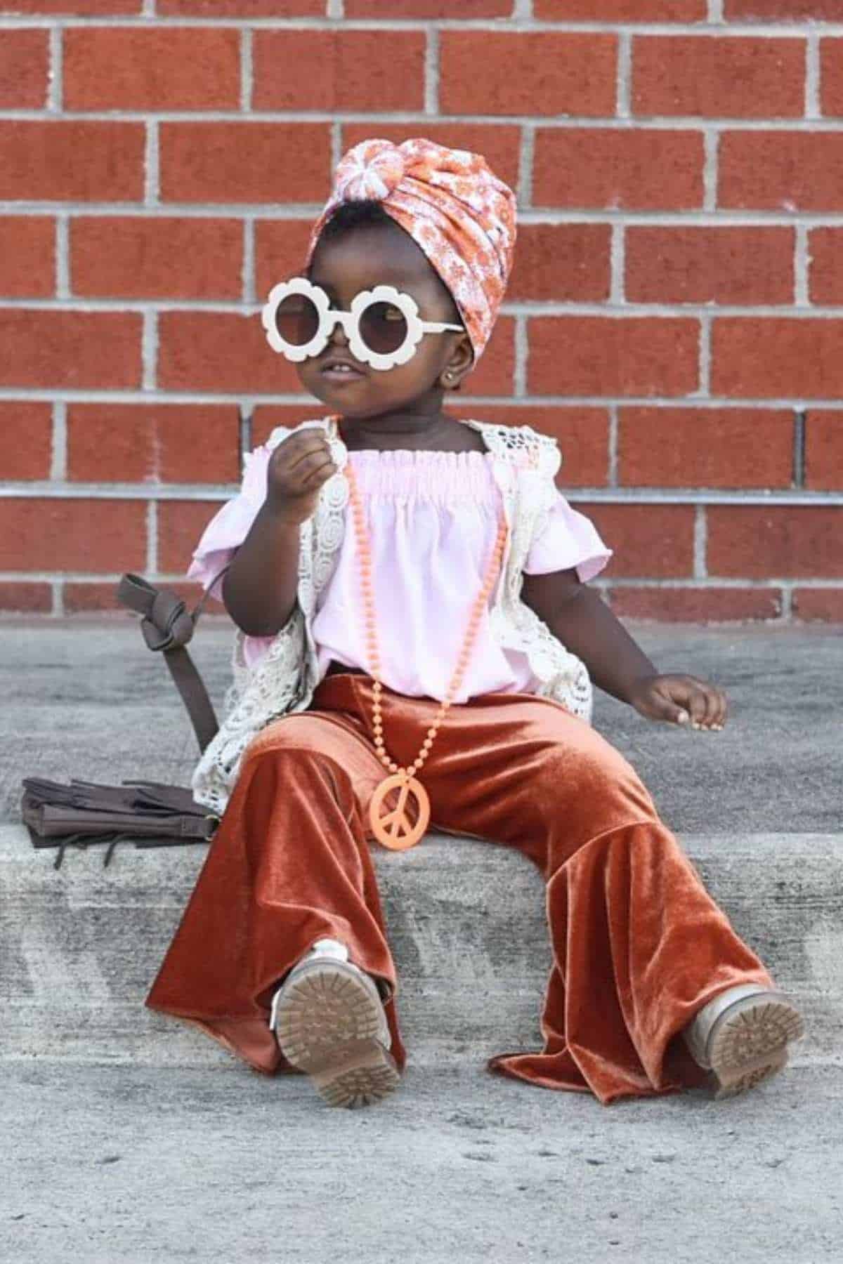 Young child sits on a concrete ledge wearing flared velvet pants, a pink top, white flower sunglasses, a headscarf, and a large peace sign necklace, with a brick wall in the background.
