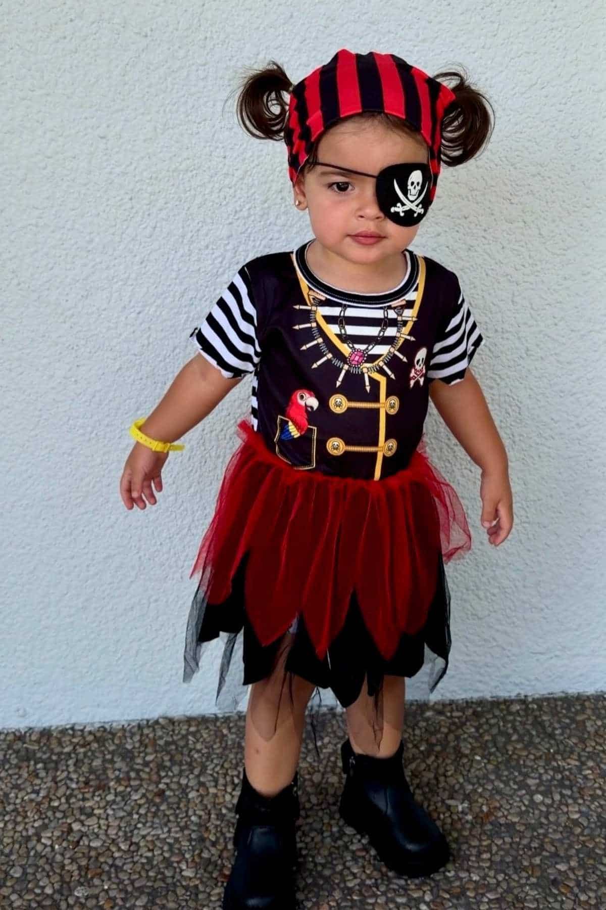 A young child dressed as a pirate, wearing a striped shirt, red-and-black skirt, eyepatch with skull and crossbones, and a matching headscarf, standing against a pale wall.