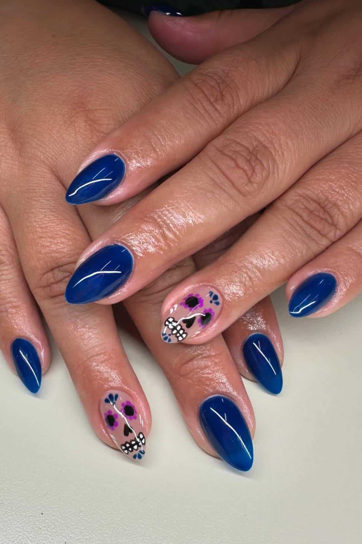 Hands with almond-shaped blue nails; two accent nails feature floral and bow designs in purple, black, and white on a nude base.