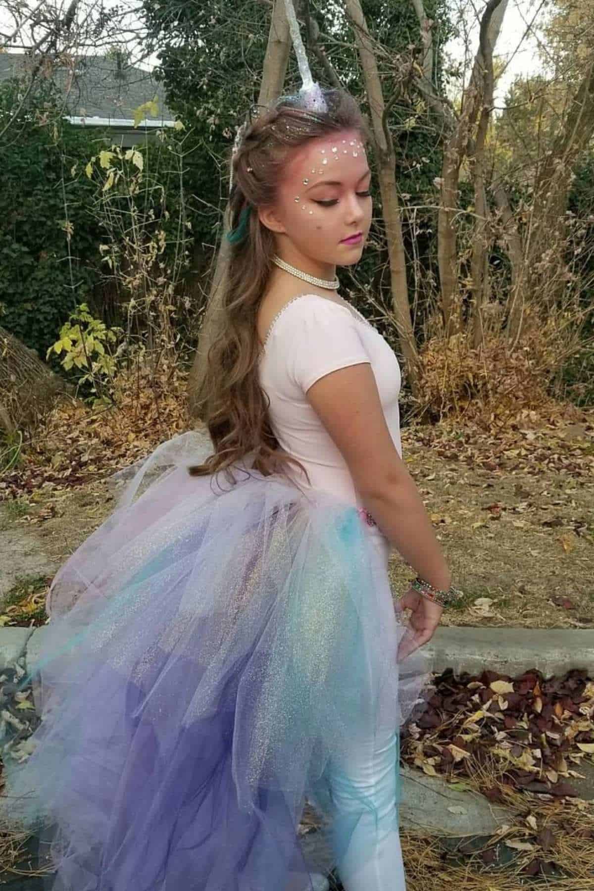 Young person in a unicorn costume with a tulle skirt and horn headband stands outdoors on a sidewalk surrounded by autumn trees and leaves.