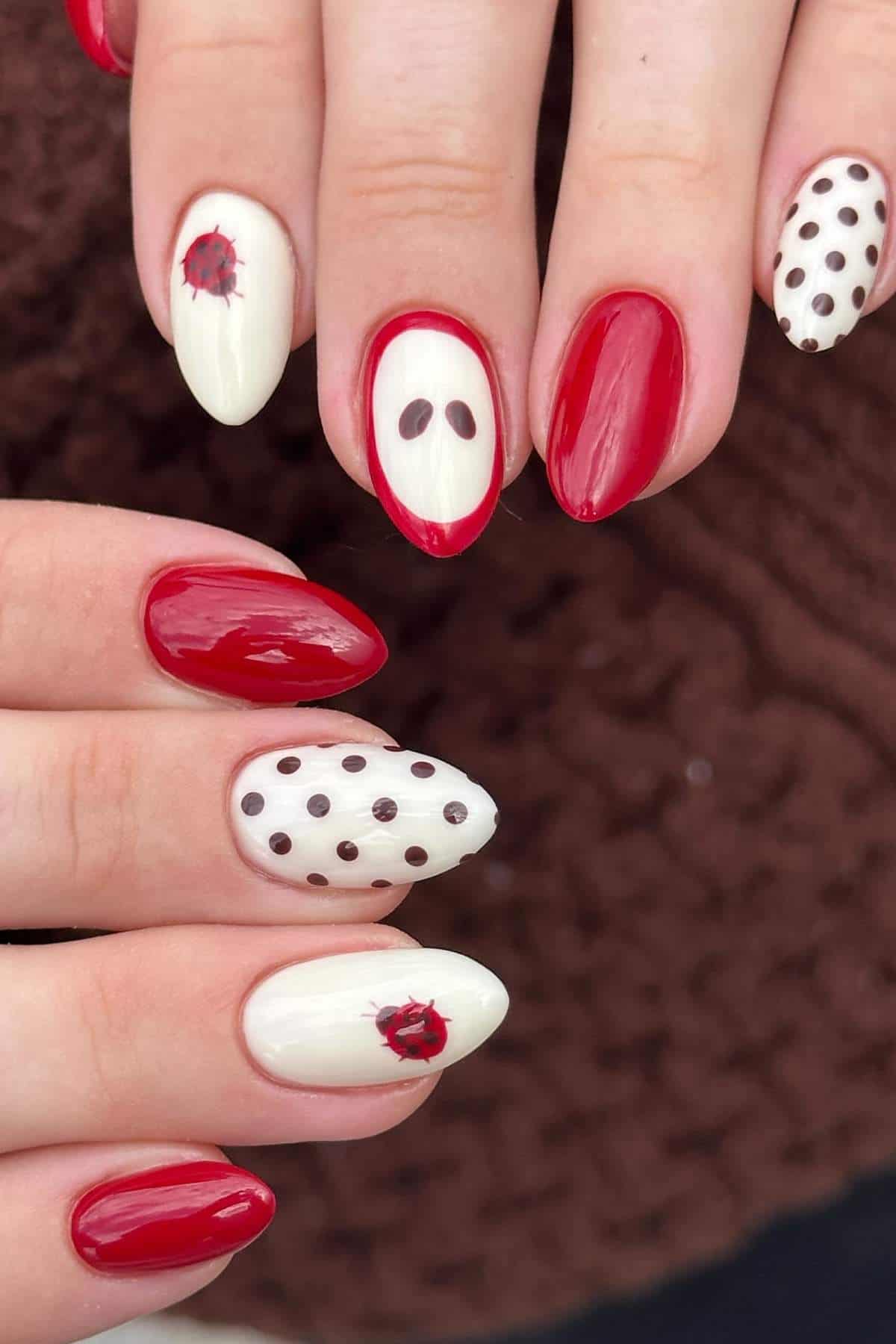 A hand with manicured nails featuring red polish, white polish with black dots, and small ladybug designs on a textured brown background.